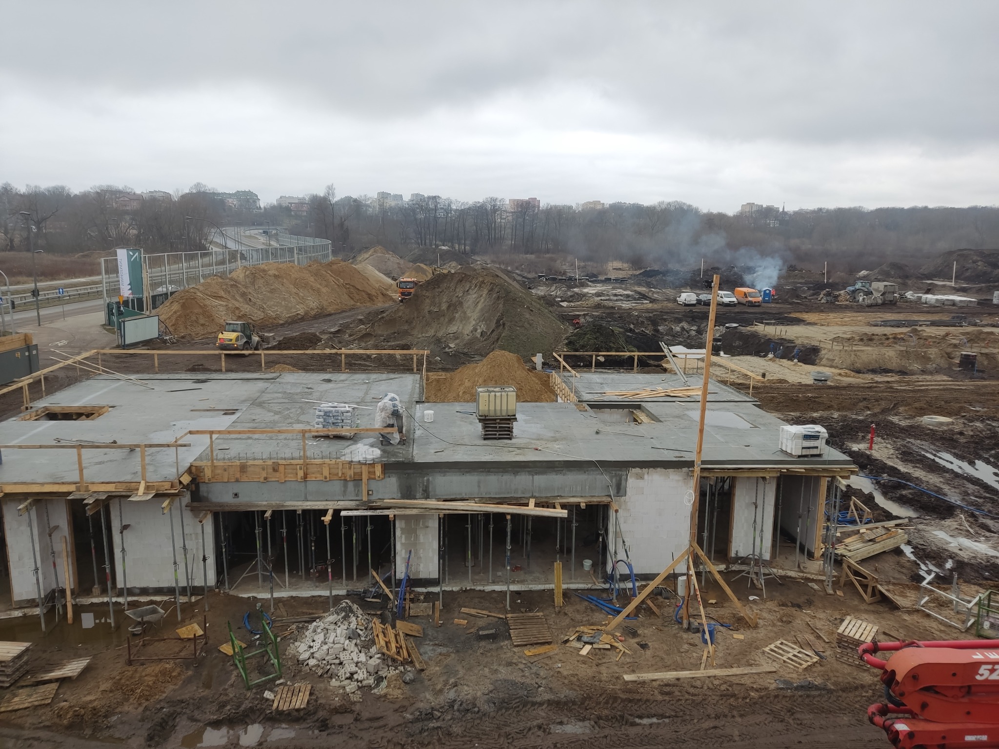 Wysoki kąt widzenia na plac budowy z widocznymi fundamentami i ścianami budynku w trakcie wznoszenia, z rozciągającym się w tle krajobrazem miejskim i hałdami ziemi.