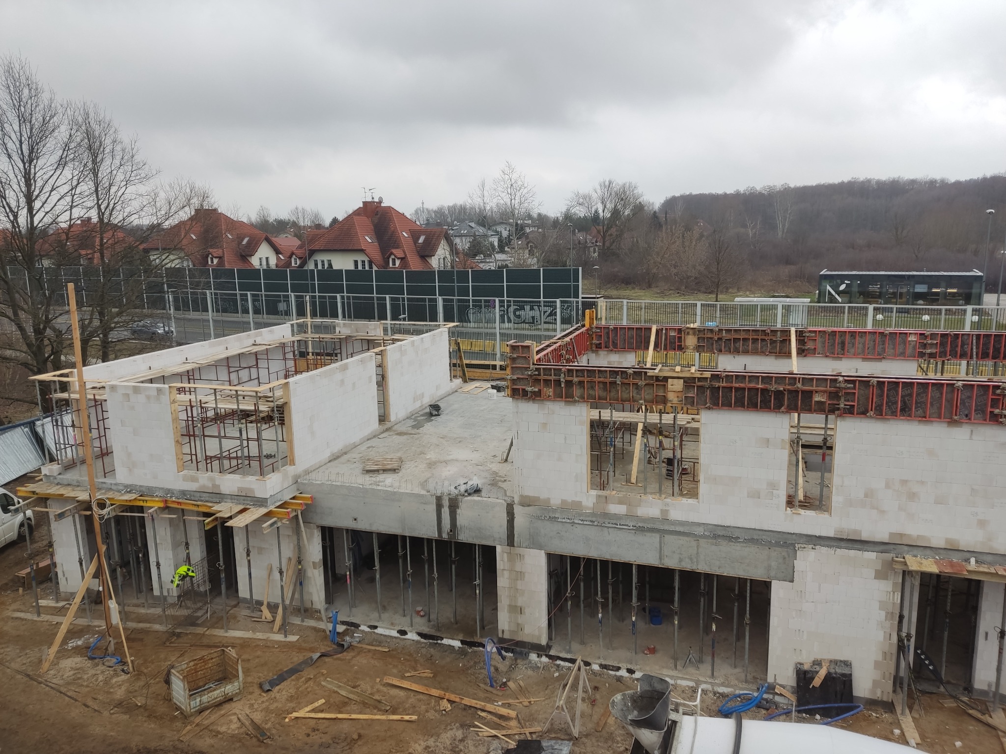 Wysoki kąt widoku na budowę domu z białych bloków, widoczne szalunki okienne, podpory stalowe i częściowo wylany strop, na drugim planie zabudowa mieszkalna i drzewa.