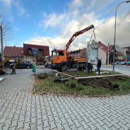 SG-USŁUGI - Montaż tablicy informacyjnej przy użyciu dźwigu samochodowego, obok wykopu i minikoparki CAT, na tle zabudowy miejskiej.