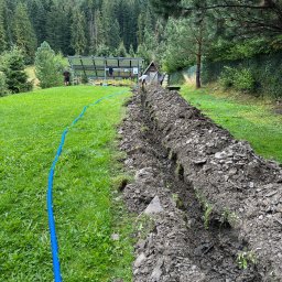 SG-USŁUGI - Wykopany rów w trawie z niebieską rurą drenarską, widoczne panele słoneczne w tle i zalesione wzgórza.