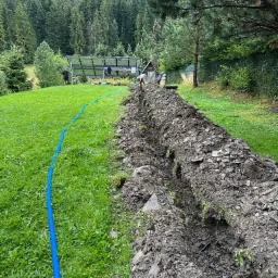 Wykopany rów w trawie z niebieską rurą drenarską, widoczne panele słoneczne w tle i zalesione wzgórza.