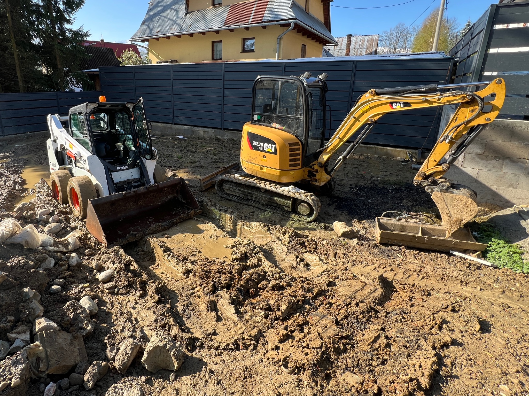 Dwa urządzenia budowlane: biała miniładowarka Bobcat i żółta minikoparka CAT 302.7D, pracują na błotnistym terenie budowy z widocznymi kamieniami, ogrodzeniem i budynkiem w tle.