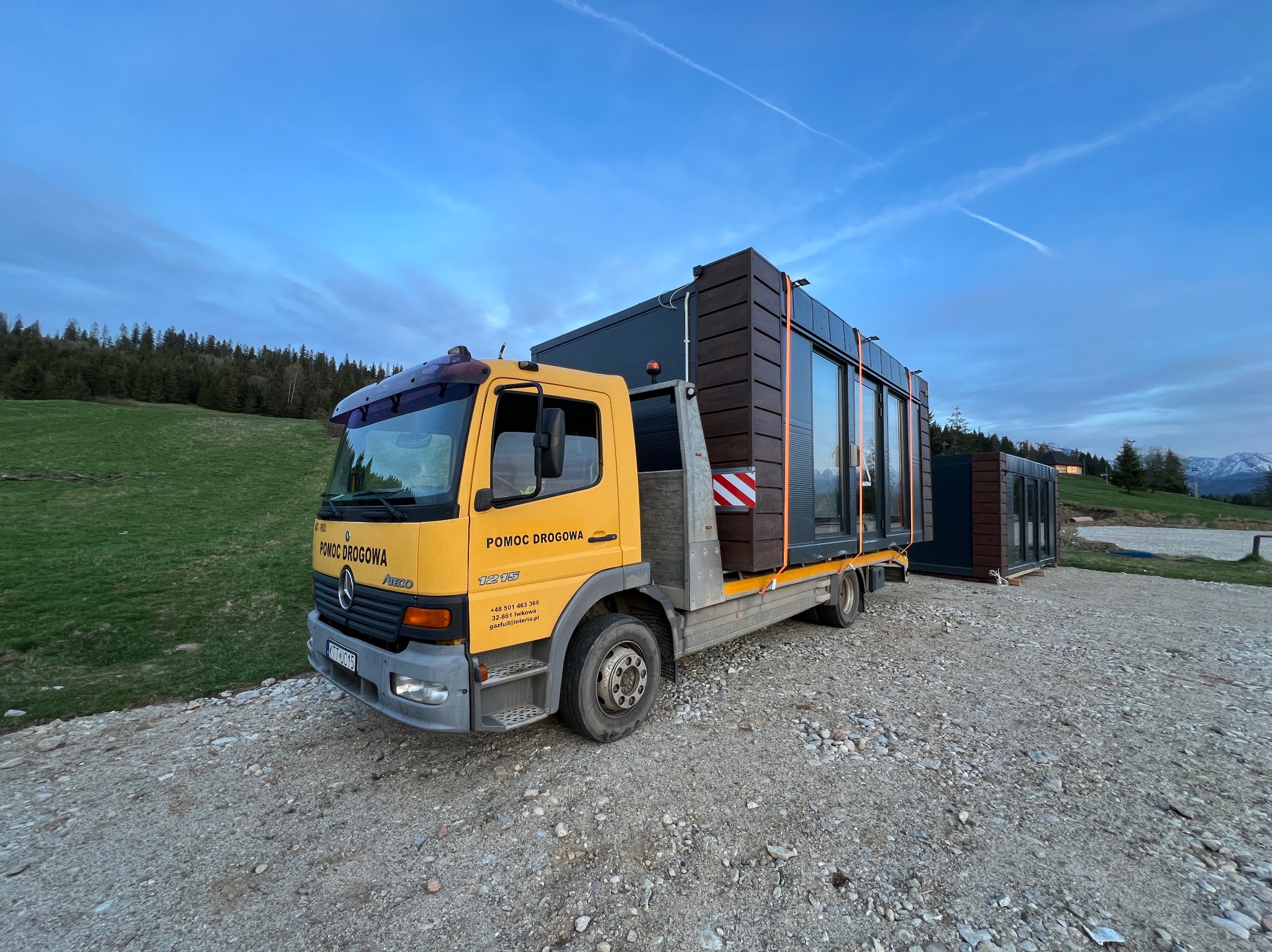 Żółta laweta Mercedes-Benz Pomoc Drogowa przewozi modułowy dom z ciemnego drewna i dużymi oknami na tle zielonych wzgórz i błękitnego nieba z lekkimi chmurami.