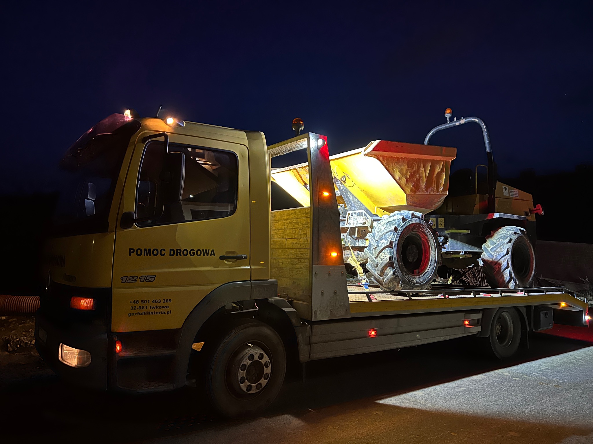Żółta laweta 'Pomoc Drogowa' przewozi mini wywrotkę budowlaną w nocnych warunkach, widoczne światła ostrzegawcze i tablice rejestracyjne.
