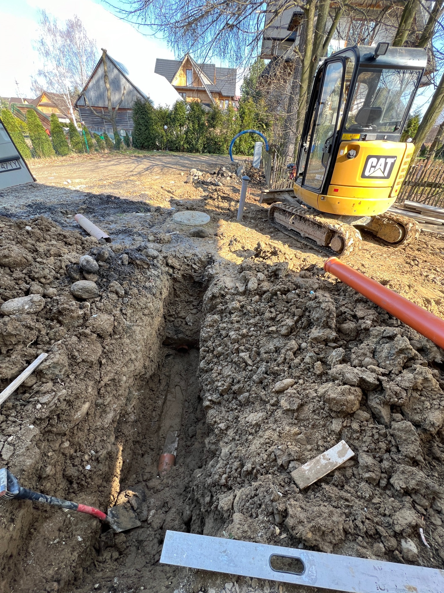 Wykop liniowy w ziemi, widoczne rury, poziomica, łopata oraz mała koparka CAT w tle, domy w oddali.