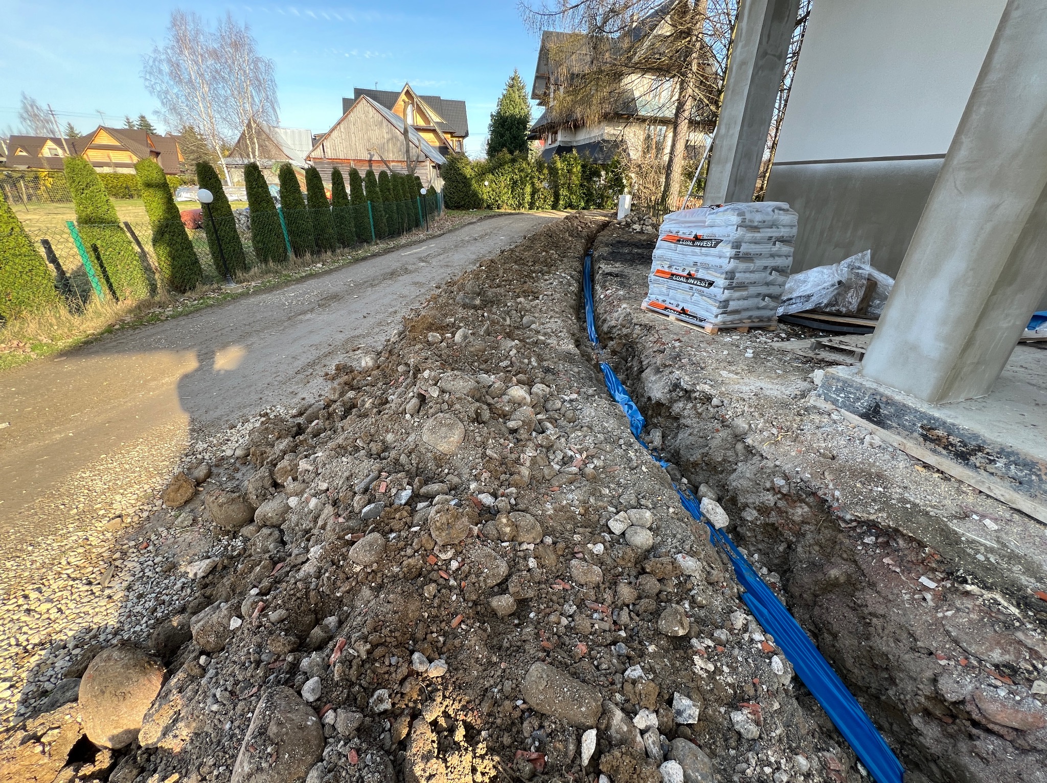 Wykopany rów z niebieską rurą w ziemi, obok sterta worków z zaprawą, domy w tle.