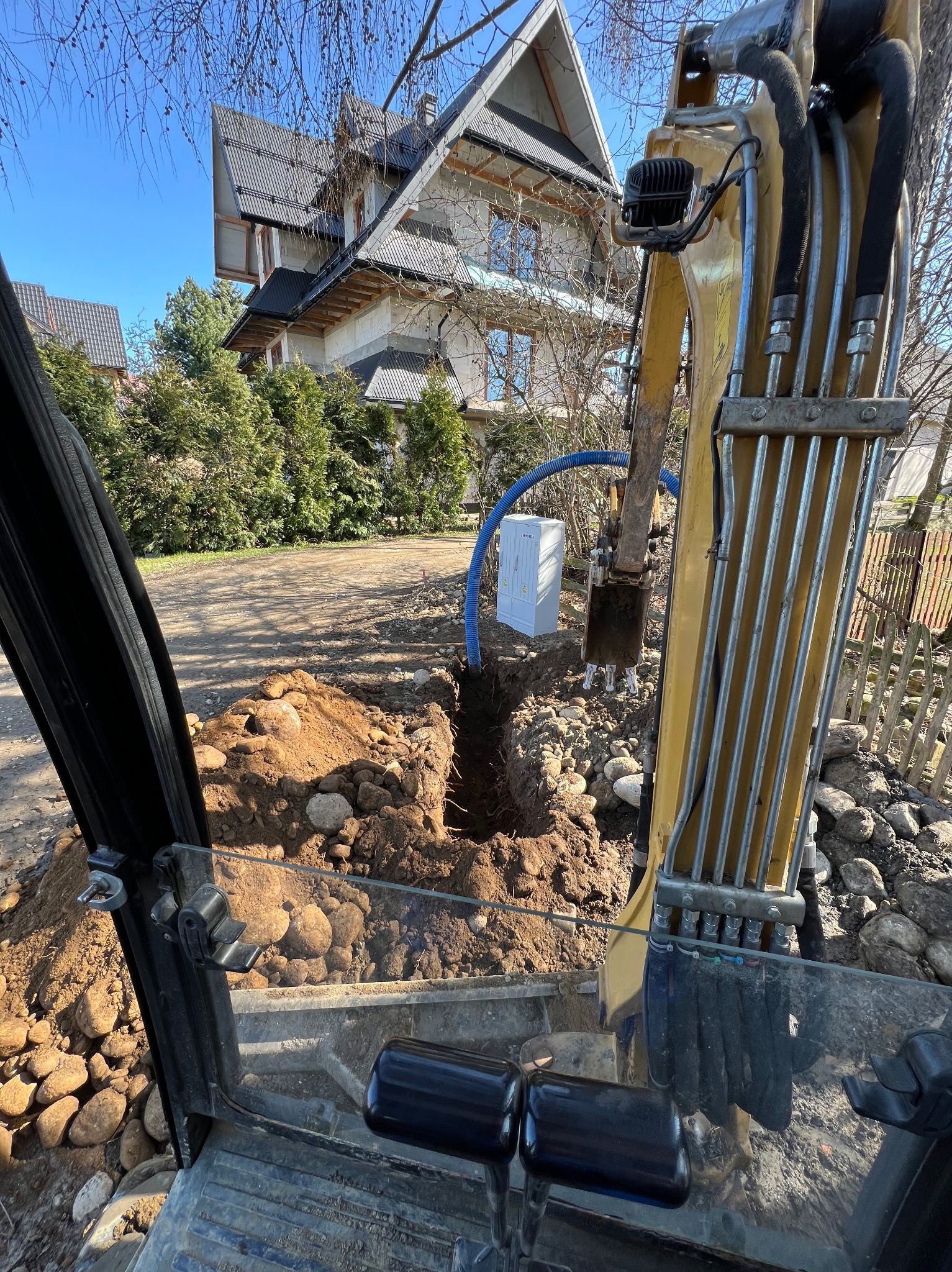 Wąski wykop w ziemi z dużą ilością kamieni, widziany z kabiny żółtej koparki, w tle biała skrzynka i dom w budowie.