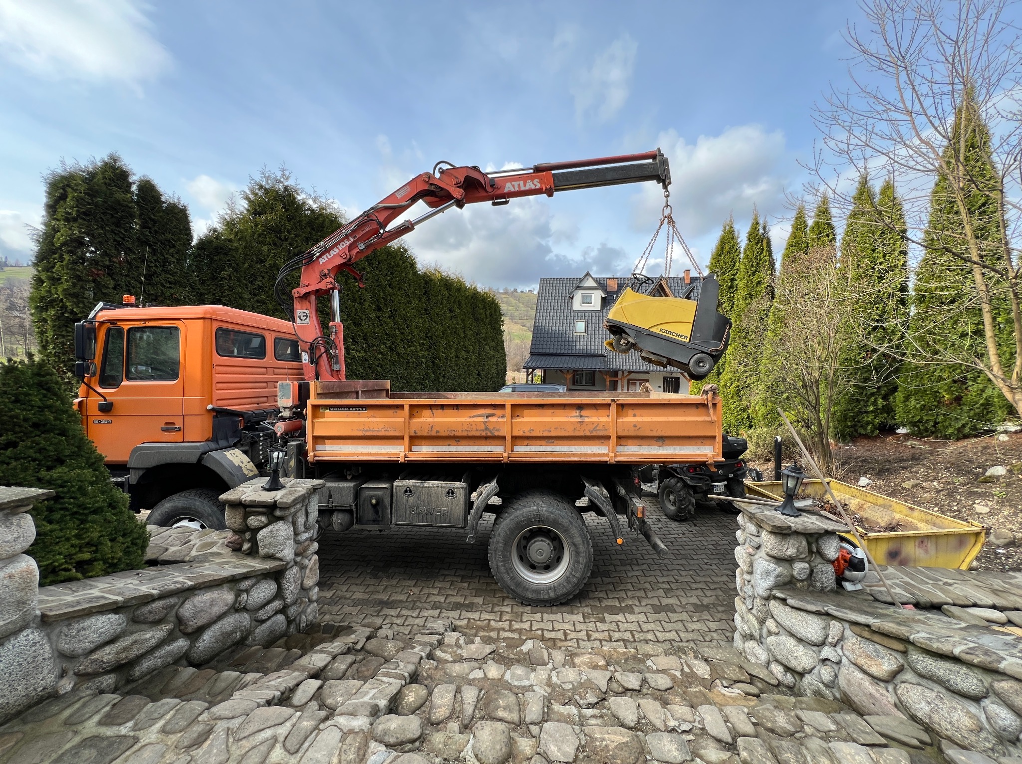 Pomarańczowy samochód ciężarowy z dźwigiem Atlas podnosi żółtą zamiatarkę Karcher nad skrzynią ładunkową na tle domu i iglaków.