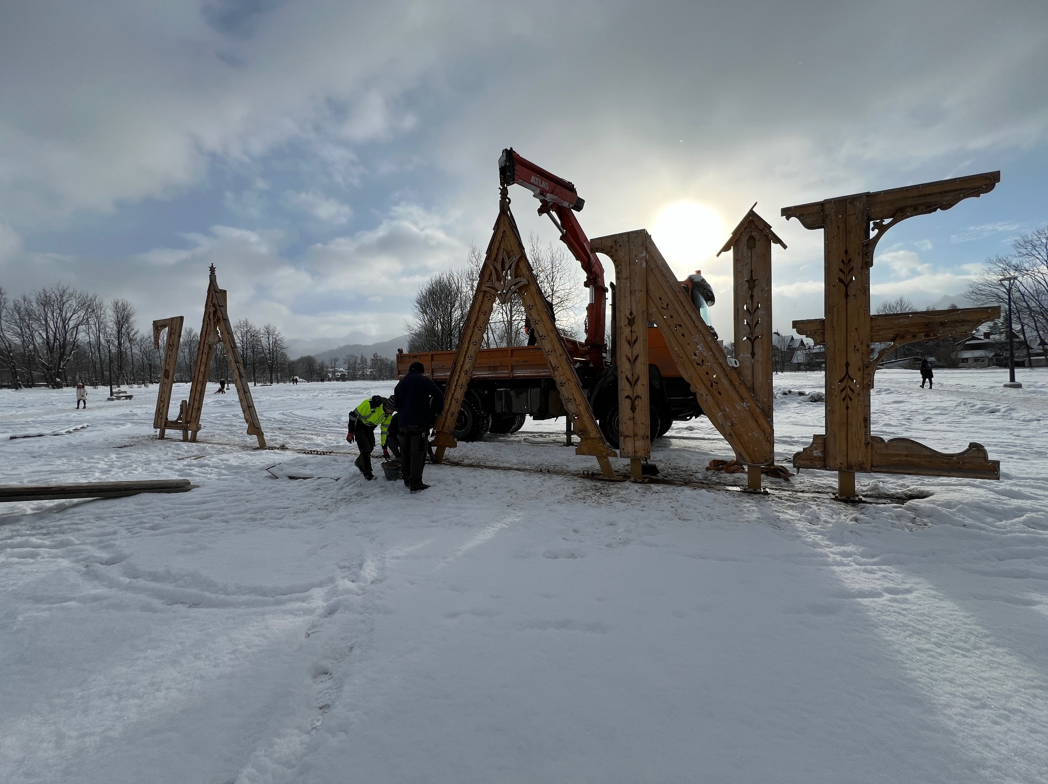 Montaż dużych, drewnianych liter dekoracyjnych na śniegu przy użyciu dźwigu samochodowego, widoczni robotnicy w odblaskowych kamizelkach.