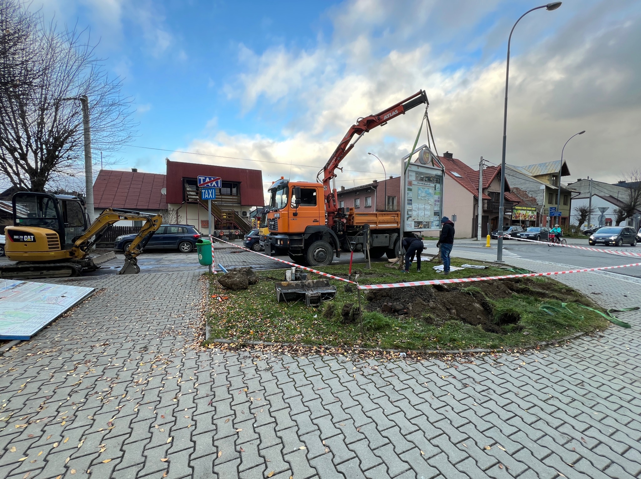 Montaż tablicy informacyjnej przy użyciu dźwigu samochodowego, obok wykopu i minikoparki CAT, na tle zabudowy miejskiej.