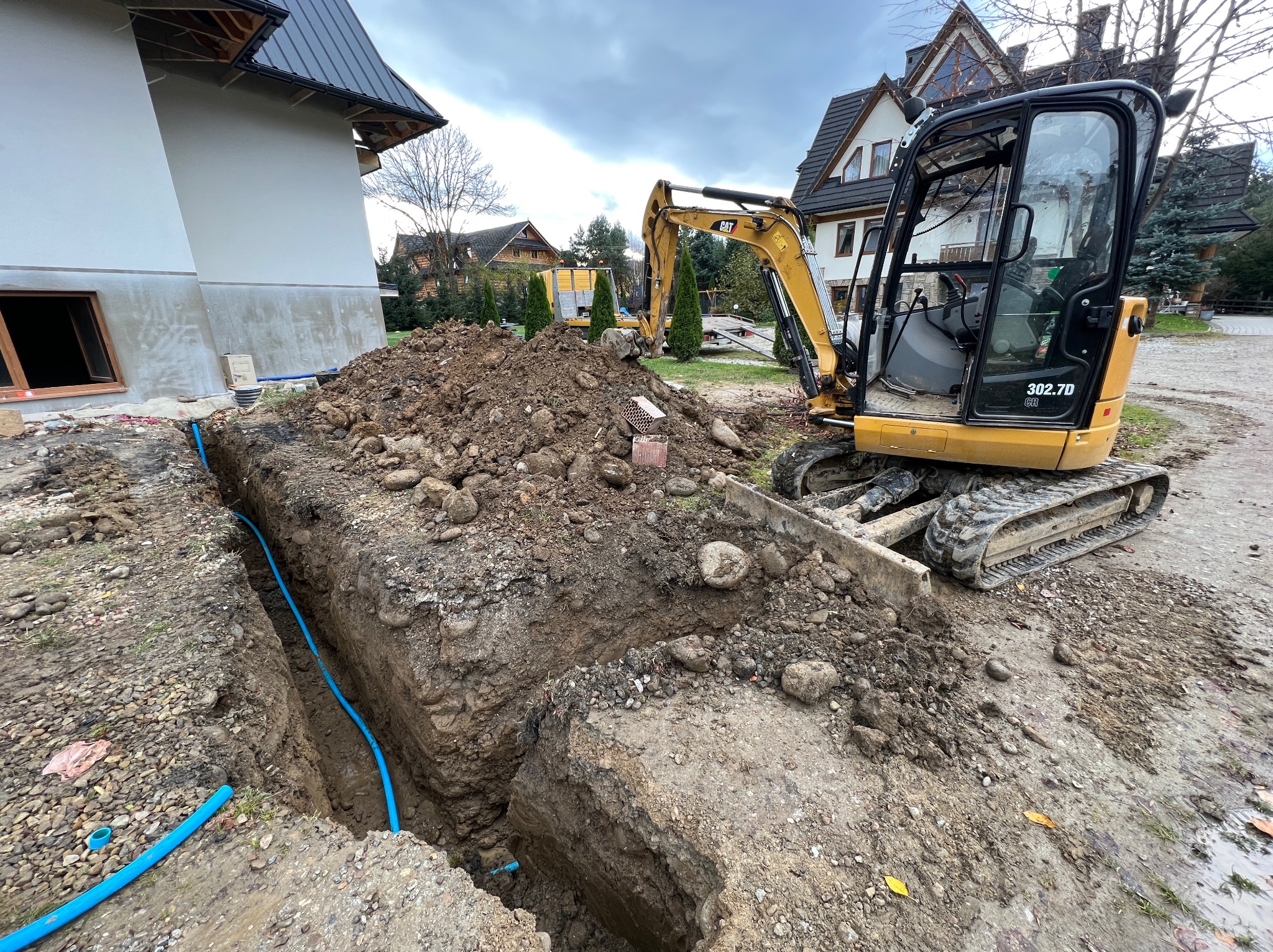 Żółta minikoparka CAT 302.7D na placu budowy, obok wykopu z niebieskimi rurami, w tle domy w stylu góralskim i koparka z lawetą.