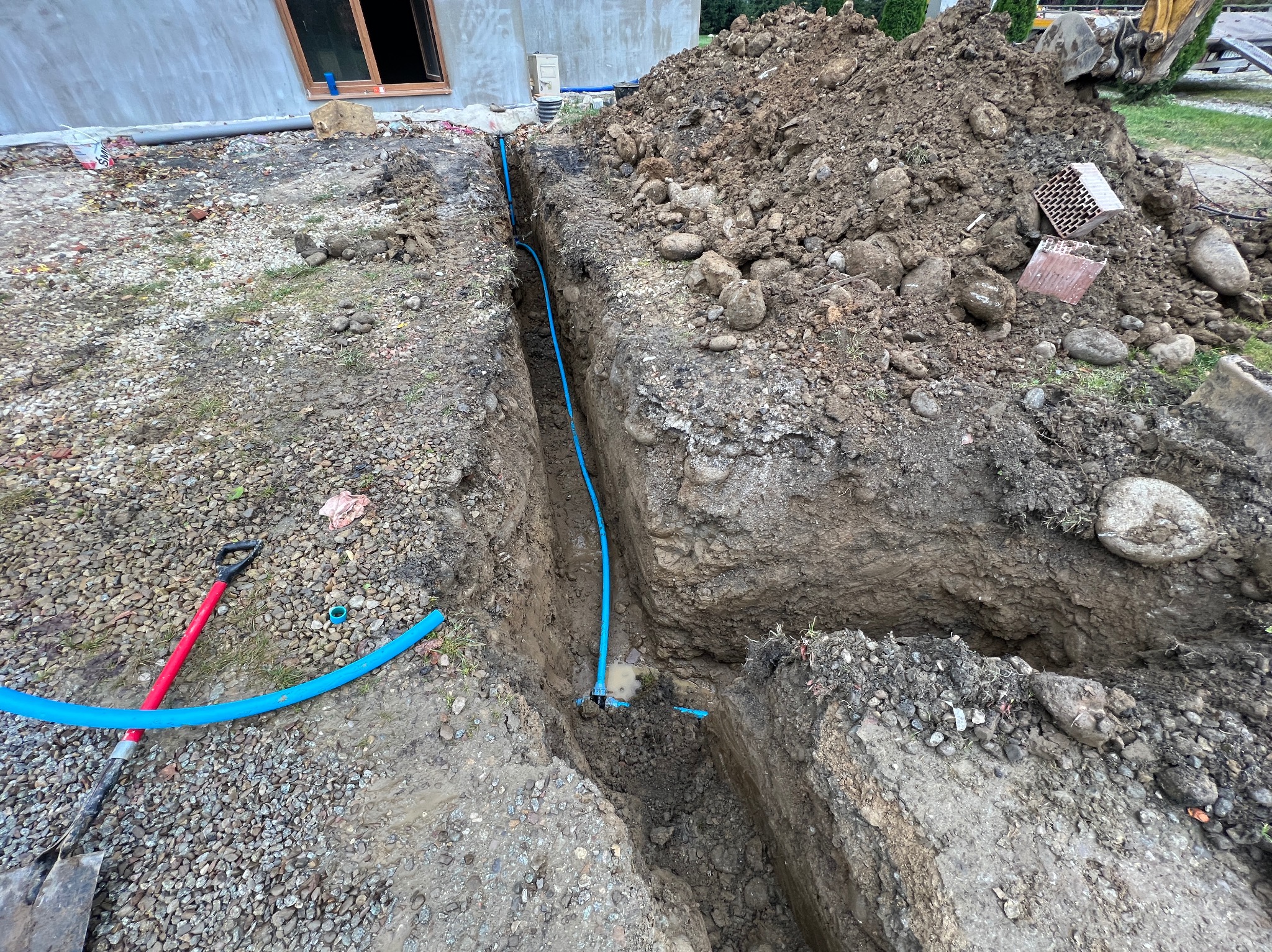Wykopany rów z ułożoną niebieską rurą, widoczna sterta ziemi i kamieni, w tle budynek w trakcie budowy i fragment koparki.
