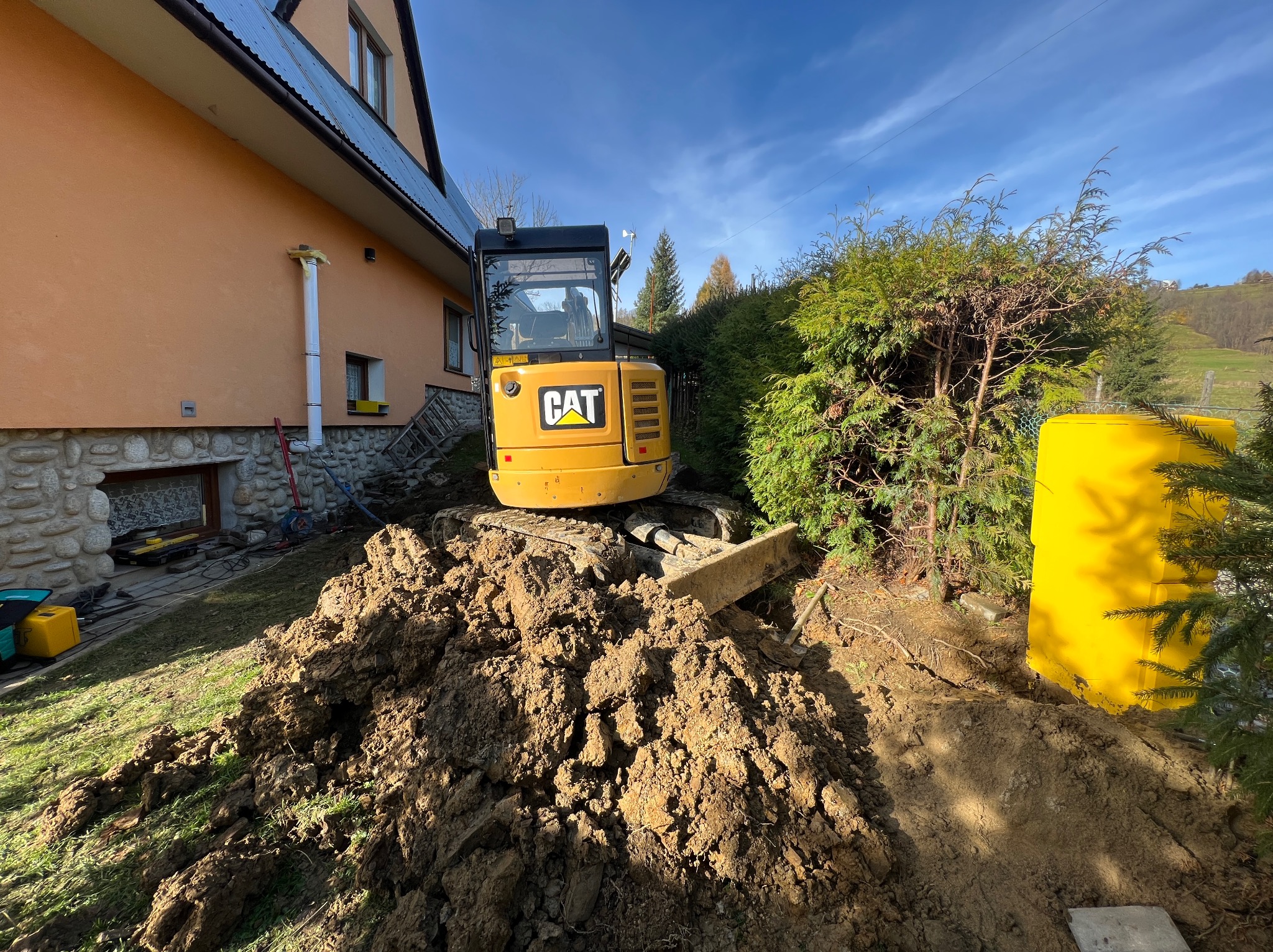 Żółta mini koparka CAT na nierównym terenie z pryzmą ziemi, obok budynku mieszkalnego i zielonych krzewów w słoneczny dzień.