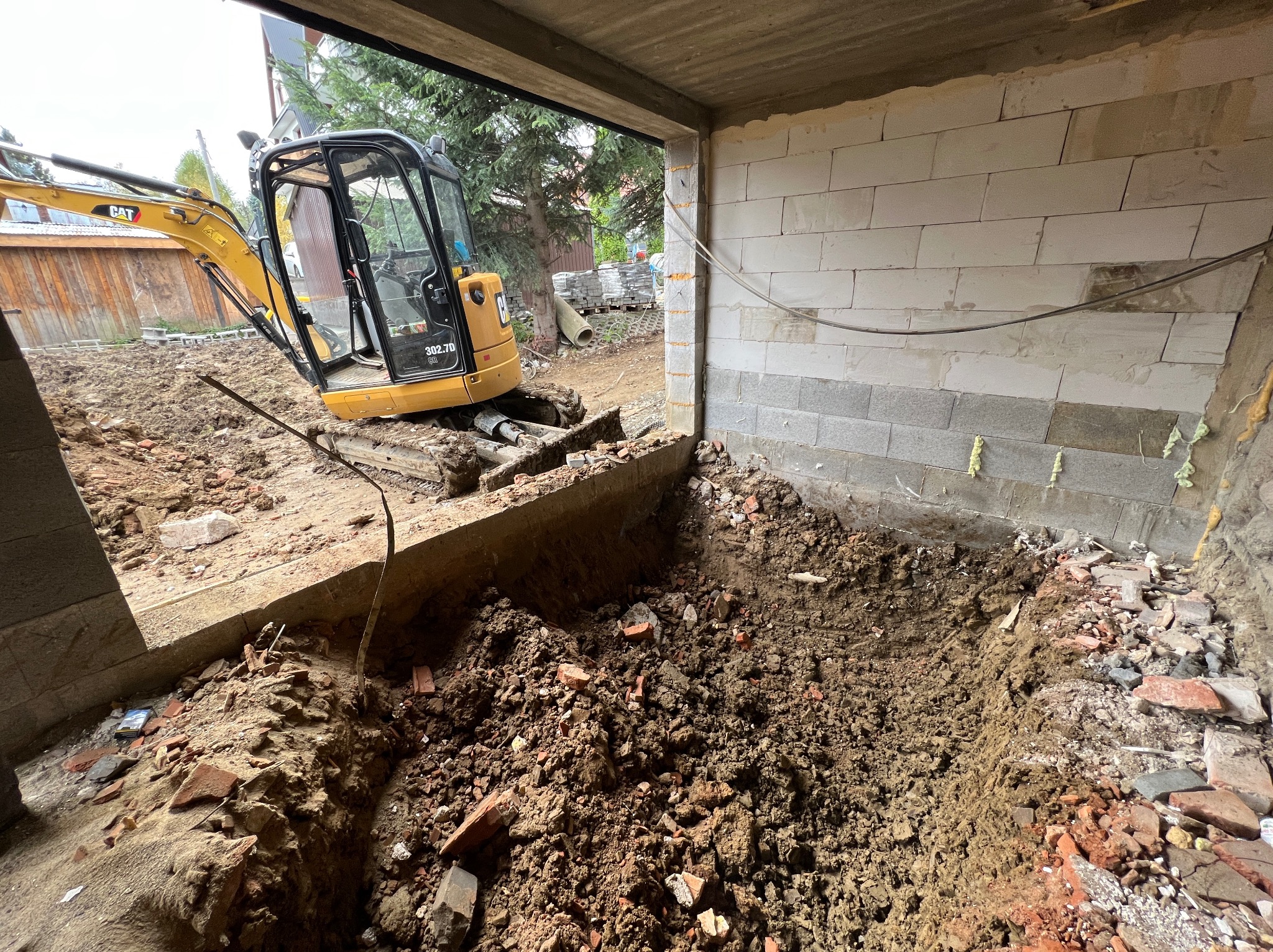 Żółta mini koparka CAT 302.7D w trakcie prac ziemnych wewnątrz budynku w stanie surowym, widoczny wykop i fragmenty cegieł.