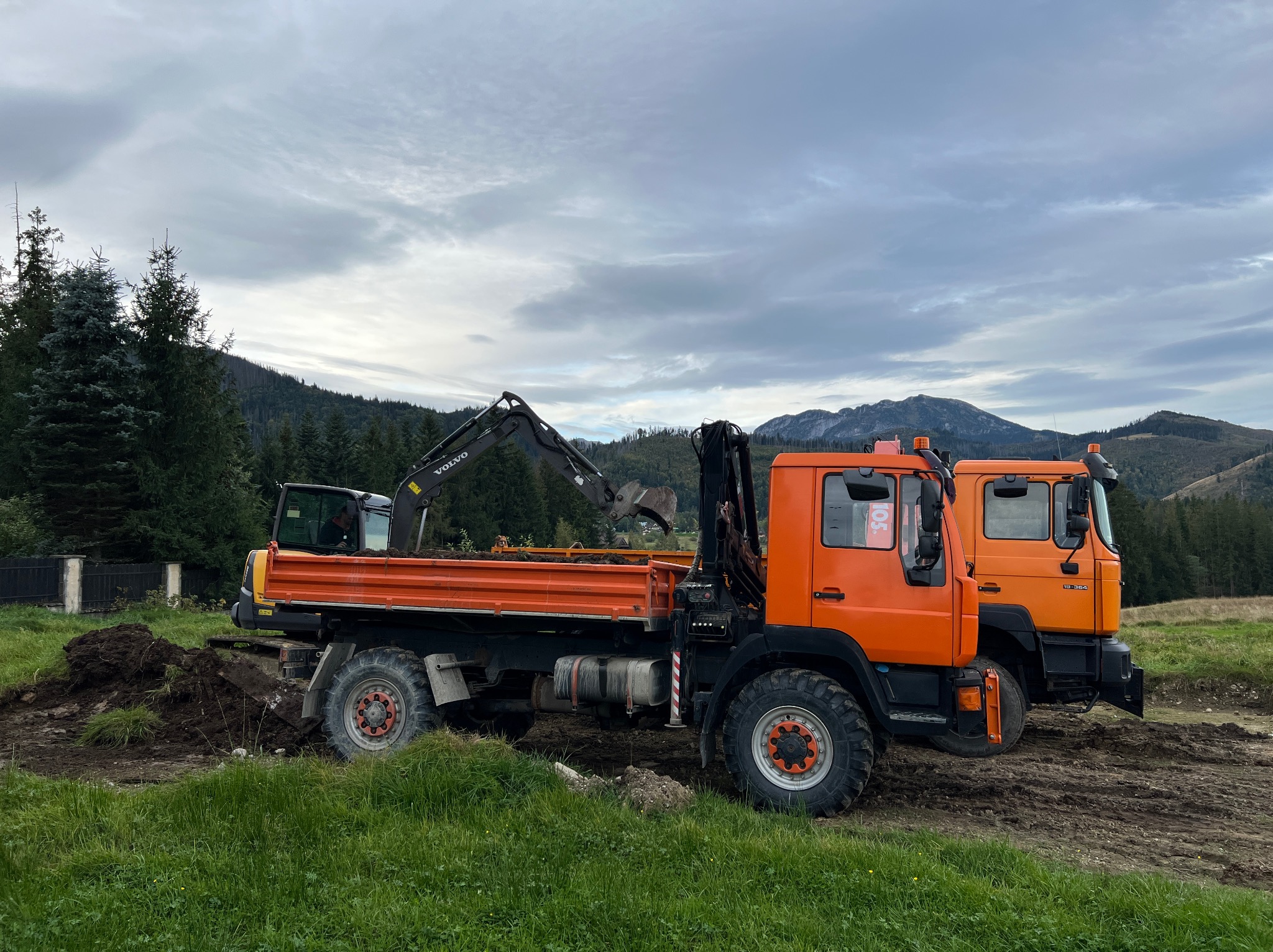 Pomarańczowa ciężarówka z koparką Volvo na skrzyni ładunkowej, obok zaparkowana druga ciężarówka, w tle góry i pochmurne niebo.