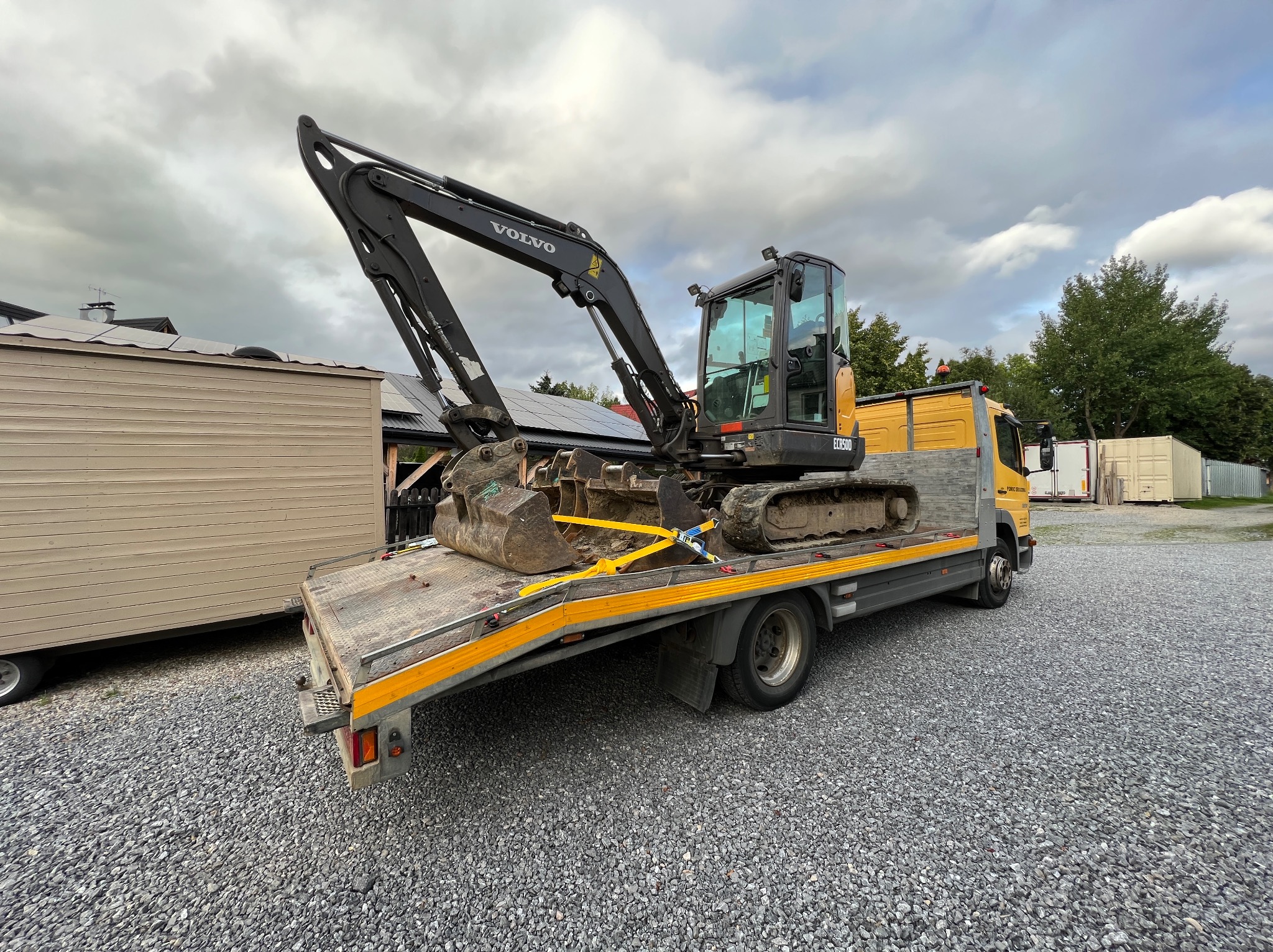 Żółta laweta z czarną mini koparką Volvo zabezpieczoną pasami transportowymi, na żwirowym podłożu, w tle budynek z panelami słonecznymi i pochmurne niebo.