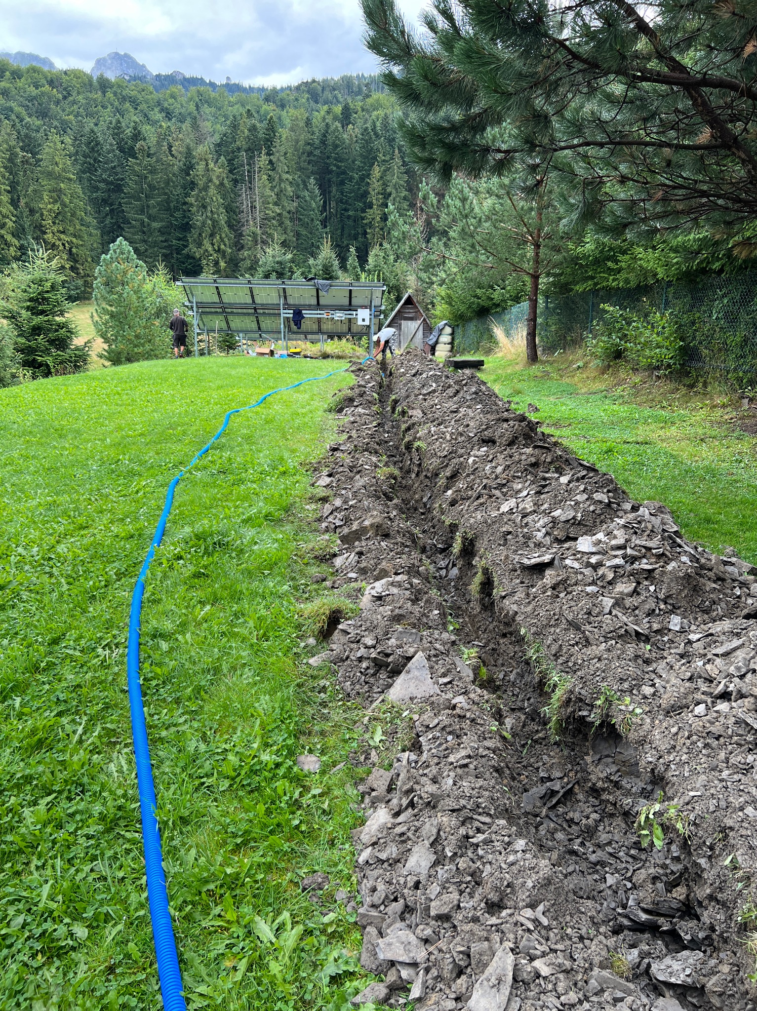 Wykopany rów w trawie z niebieską rurą drenarską, widoczne panele słoneczne w tle i zalesione wzgórza.
