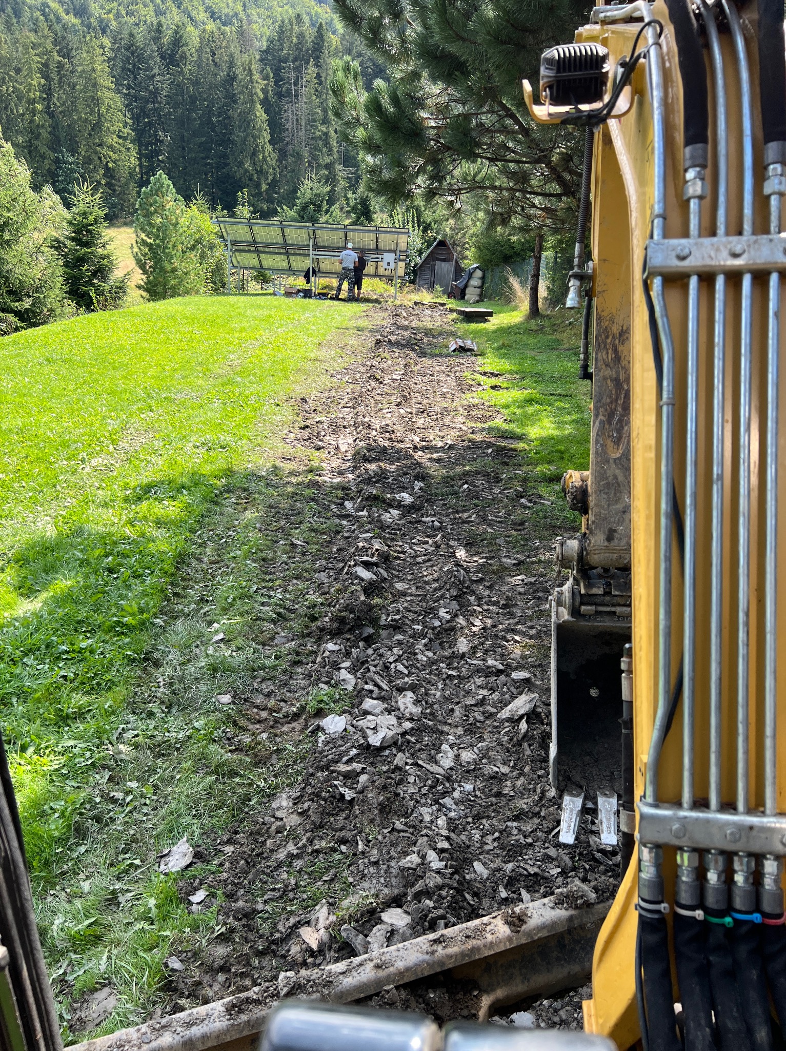 Żółta koparka na skraju wykopu, w tle panele słoneczne i zielone wzgórze.