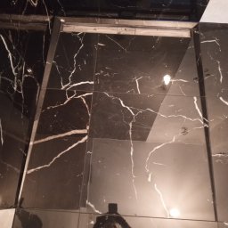 Maciej Flaga Żurawie - Błyszcząca, czarna podłoga wyłożona płytkami imitującymi marmur z białymi żyłami, widoczny fragment progu i odbicie fotografa w płytkach.