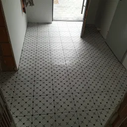 Ułożona podłoga z białych płytek z czarnymi wzorami w kształcie rombów i gwiazdek, widok na wejście z szarymi płytami chodnikowymi na zewnątrz, surowy mur z cegieł po lewej stronie.