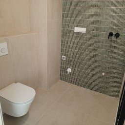 Maciej Flaga Żurawie - Wykończona łazienka z beżowymi płytkami podłogowymi i ściennymi, wisząca toaleta z przyciskiem spłukującym, ściana z zielonej mozaiki pionowej, czarna armatura podtynkowa.