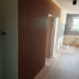 Maciej Flaga Żurawie - Nowoczesne wykończenie wnętrza: drewniane lamele na ścianie z podświetleniem LED, jasna podłoga, widoczny fragment pomieszczenia w trakcie prac wykończeniowych.