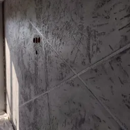 Ściana z efektem dekoracyjnym imitującym beton, podzielona listwami, z widocznymi otworami na gniazdka elektryczne, oświetlona światłem dziennym wpadającym przez okno.