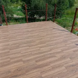 Wyłożony brązowymi panelami taras z niską, brązową barierką z czerwonym sznurkiem, widok na ogr&oacute;d z zielenią.