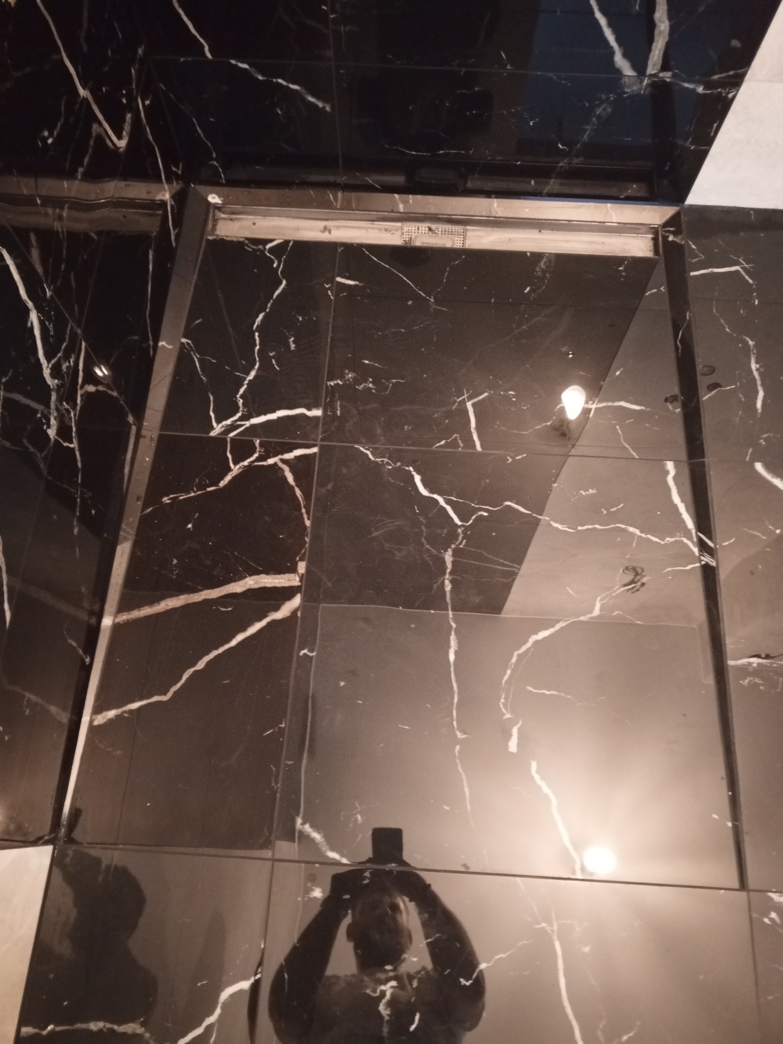 Błyszcząca, czarna podłoga wyłożona płytkami imitującymi marmur z białymi żyłami, widoczny fragment progu i odbicie fotografa w płytkach.