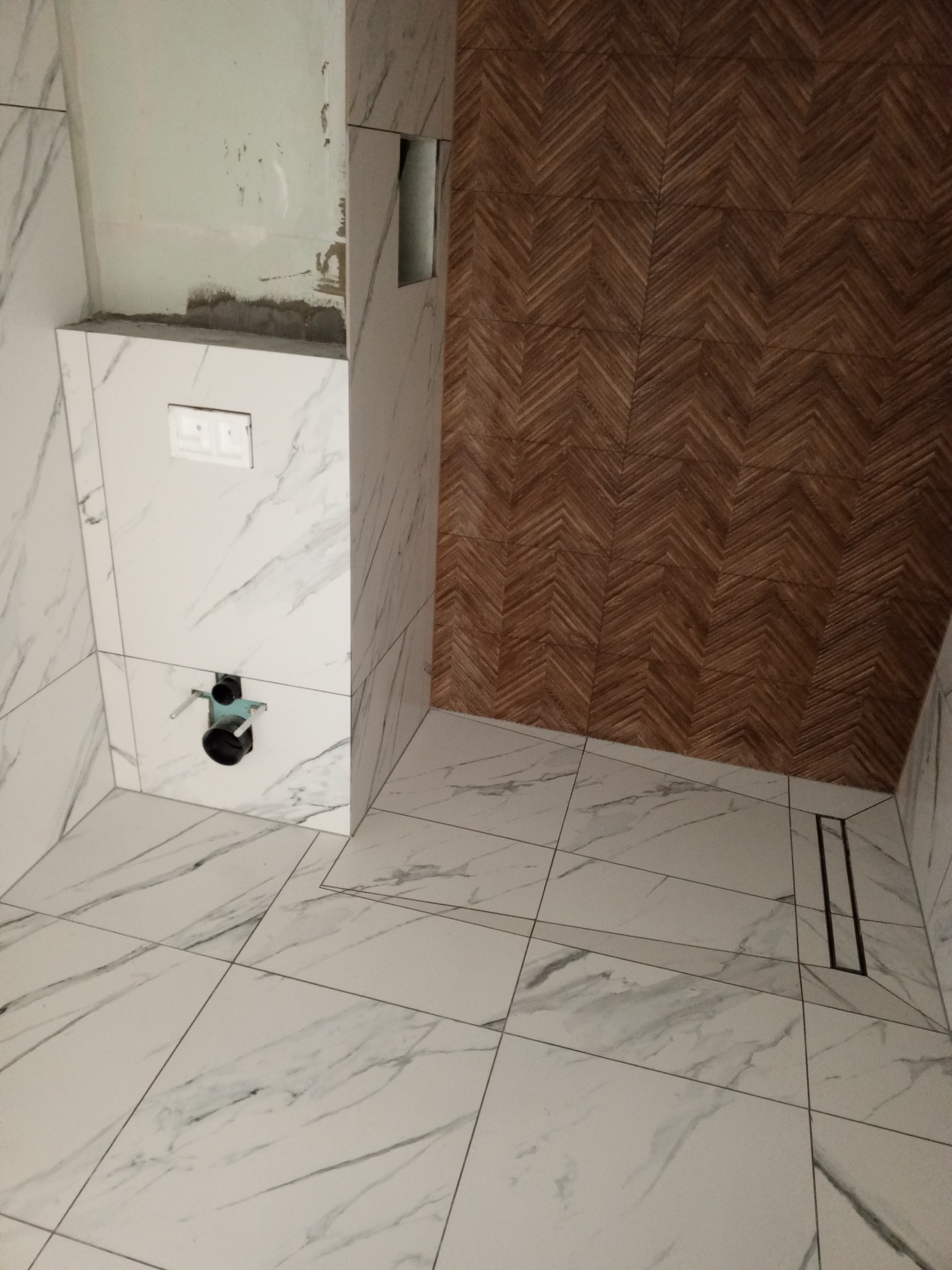 Wnętrze łazienki w trakcie prac: białe płytki imitujące marmur na podłodze i ścianach, drewnopodobne płytki ułożone w jodełkę na fragmencie ściany, widoczna instalacja podtynkowego WC i odpływ...