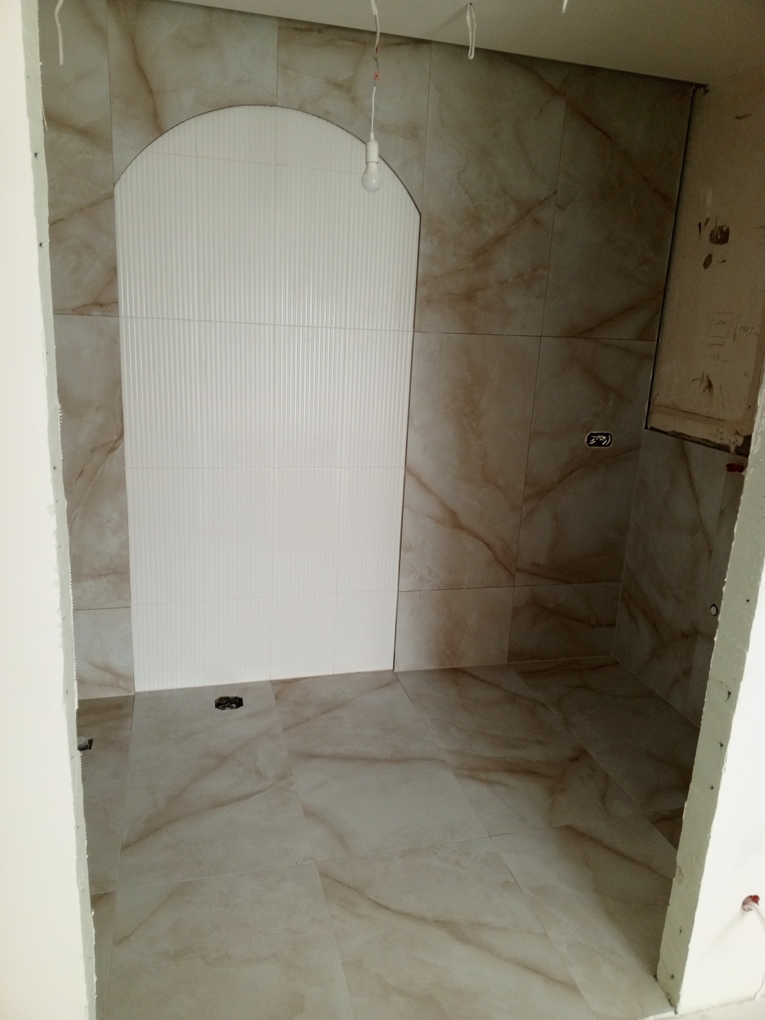 Niedokończona łazienka z beżowo-marmurkowymi płytkami na podłodze i ścianach, z wnęką wyłożoną białymi, pionowo żłobionymi płytkami w kształcie łuku, widoczna surowa żarówka i otwory na gniazdka.