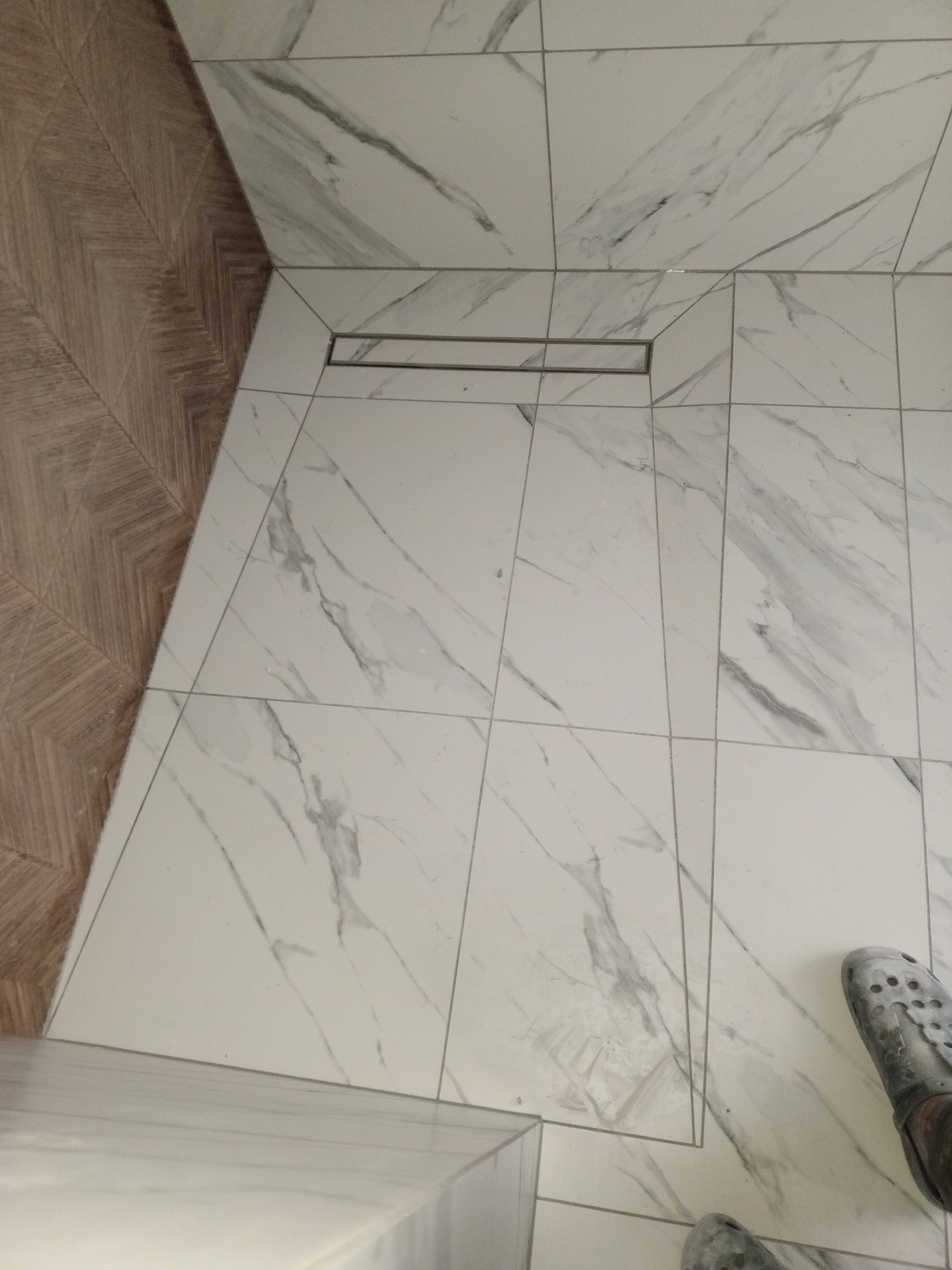 Ułożone białe płytki podłogowe z szarym marmurkowym wzorem w kabinie prysznicowej, widoczny odpływ liniowy i fragment drewnopodobnej okładziny ściennej oraz para brudnych Crocsów.