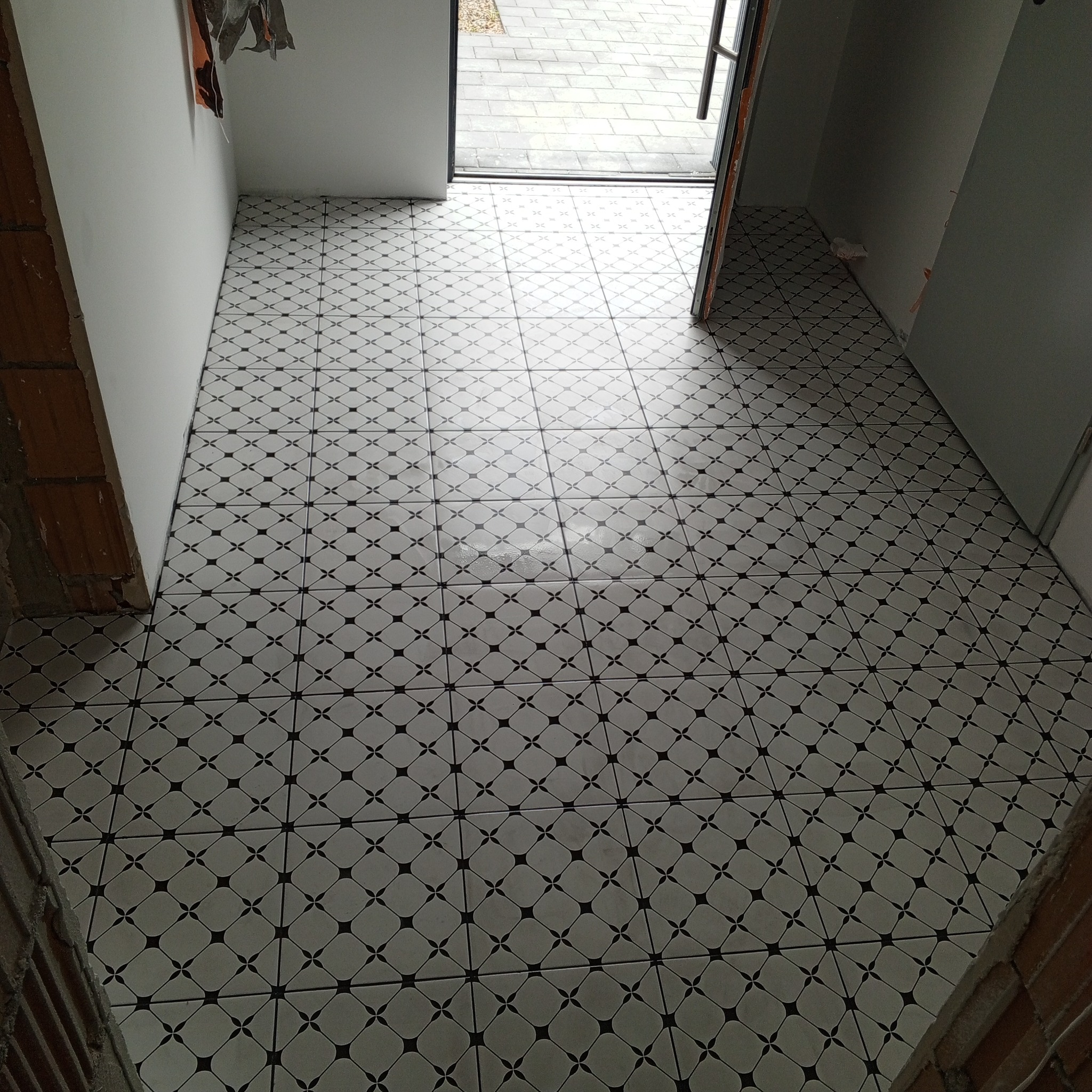 Ułożona podłoga z białych płytek z czarnymi wzorami w kształcie rombów i gwiazdek, widok na wejście z szarymi płytami chodnikowymi na zewnątrz, surowy mur z cegieł po lewej stronie.