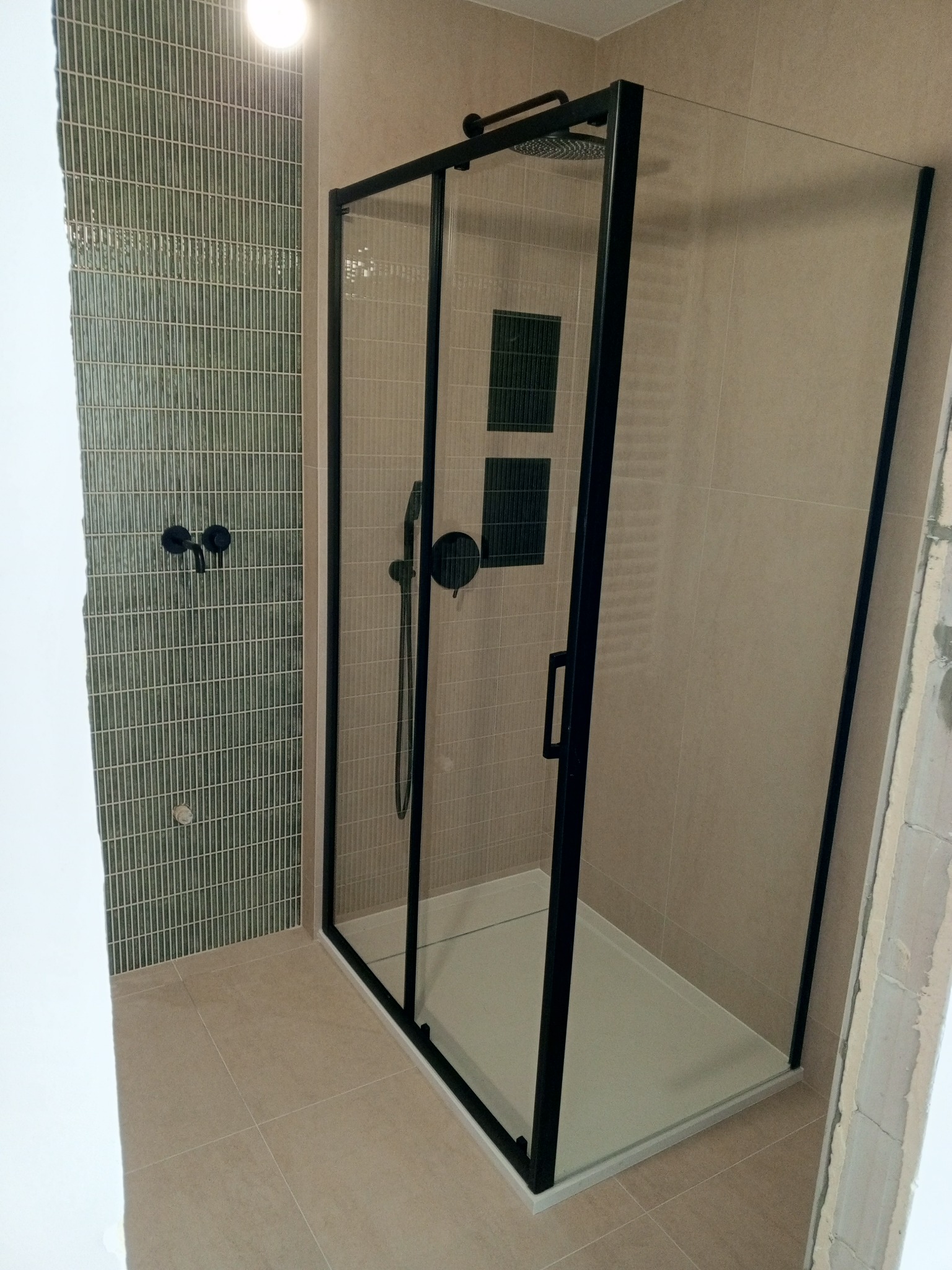 Nowoczesna kabina prysznicowa z czarną ramą i deszczownicą, zielona mozaika na ścianie, beżowe płytki podłogowe.