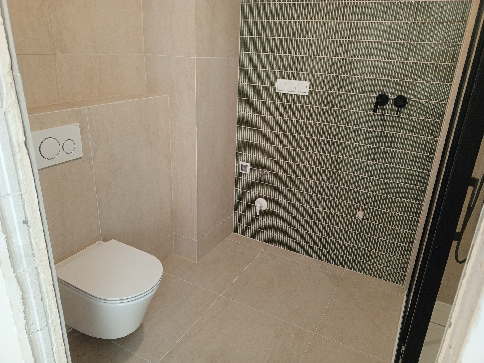 Wykończona łazienka z beżowymi płytkami podłogowymi i ściennymi, wisząca toaleta z przyciskiem spłukującym, ściana z zielonej mozaiki pionowej, czarna armatura podtynkowa.