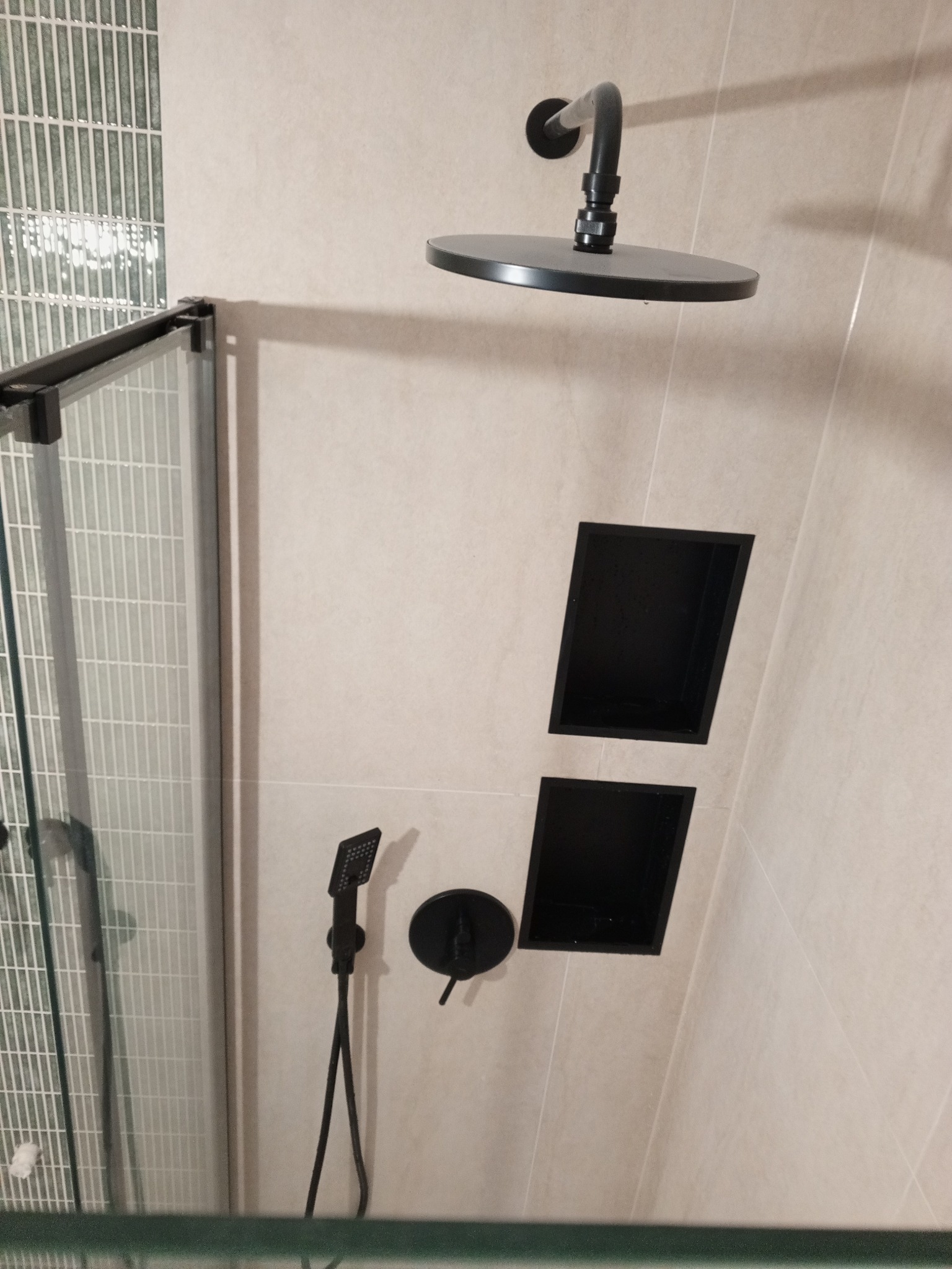 Nowoczesna kabina prysznicowa z deszczownicą w kolorze czarnym, wbudowane półki ścienne, jasne płytki ceramiczne i szklane drzwi z czarnymi ramami.