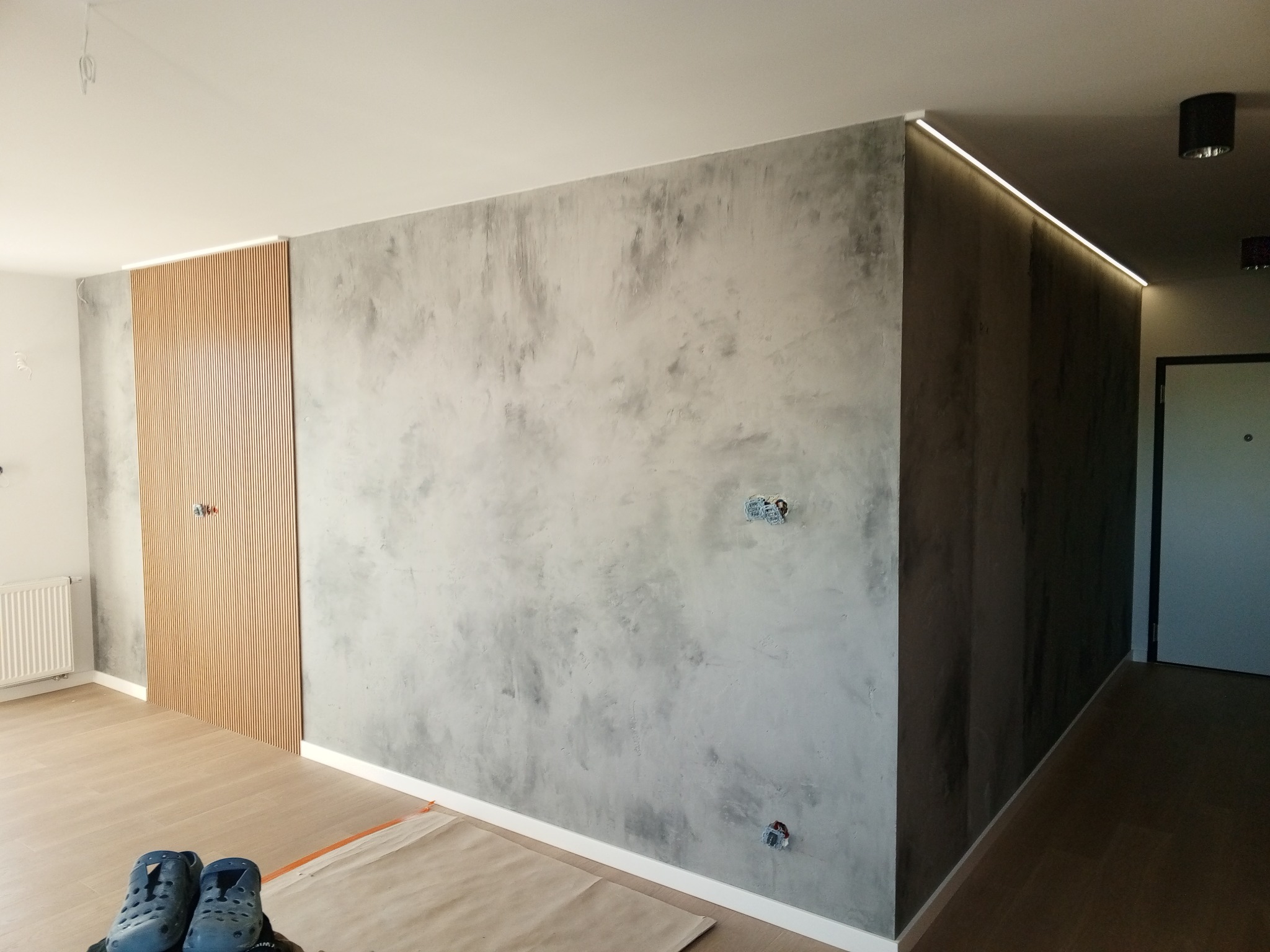 Ściana z efektem betonu architektonicznego, drewniane lamele dekoracyjne na ścianie, listwa LED podsufitowa z oświetleniem, widoczne puszki elektryczne przed montażem osprzętu.