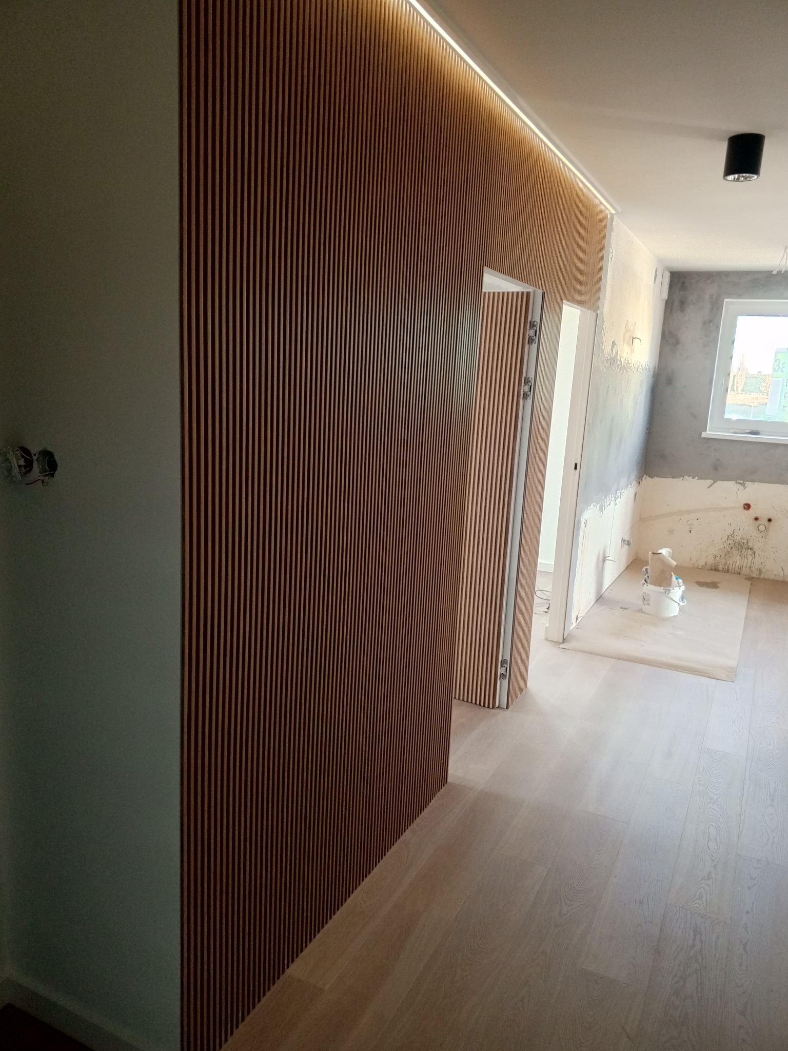 Nowoczesne wykończenie wnętrza: drewniane lamele na ścianie z podświetleniem LED, jasna podłoga, widoczny fragment pomieszczenia w trakcie prac wykończeniowych.