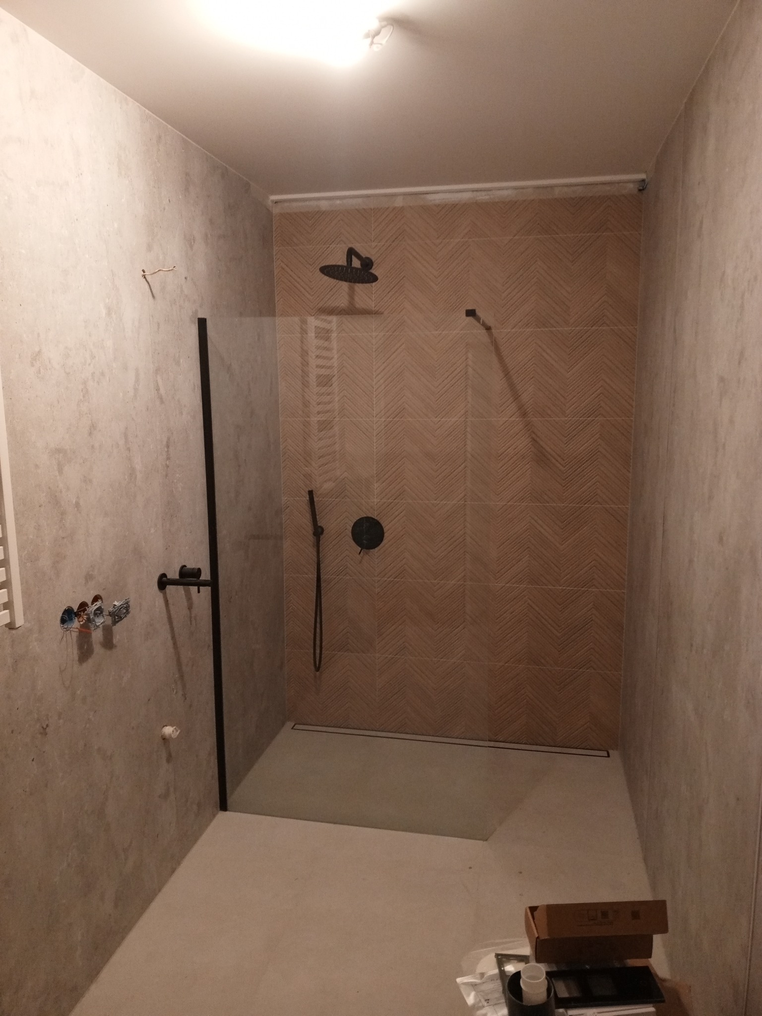 Montaż kabiny prysznicowej typu walk-in z czarną armaturą i odpływem liniowym, ściany wyłożone płytkami w geometryczny wzór, widoczne niedokończone prace instalacyjne.