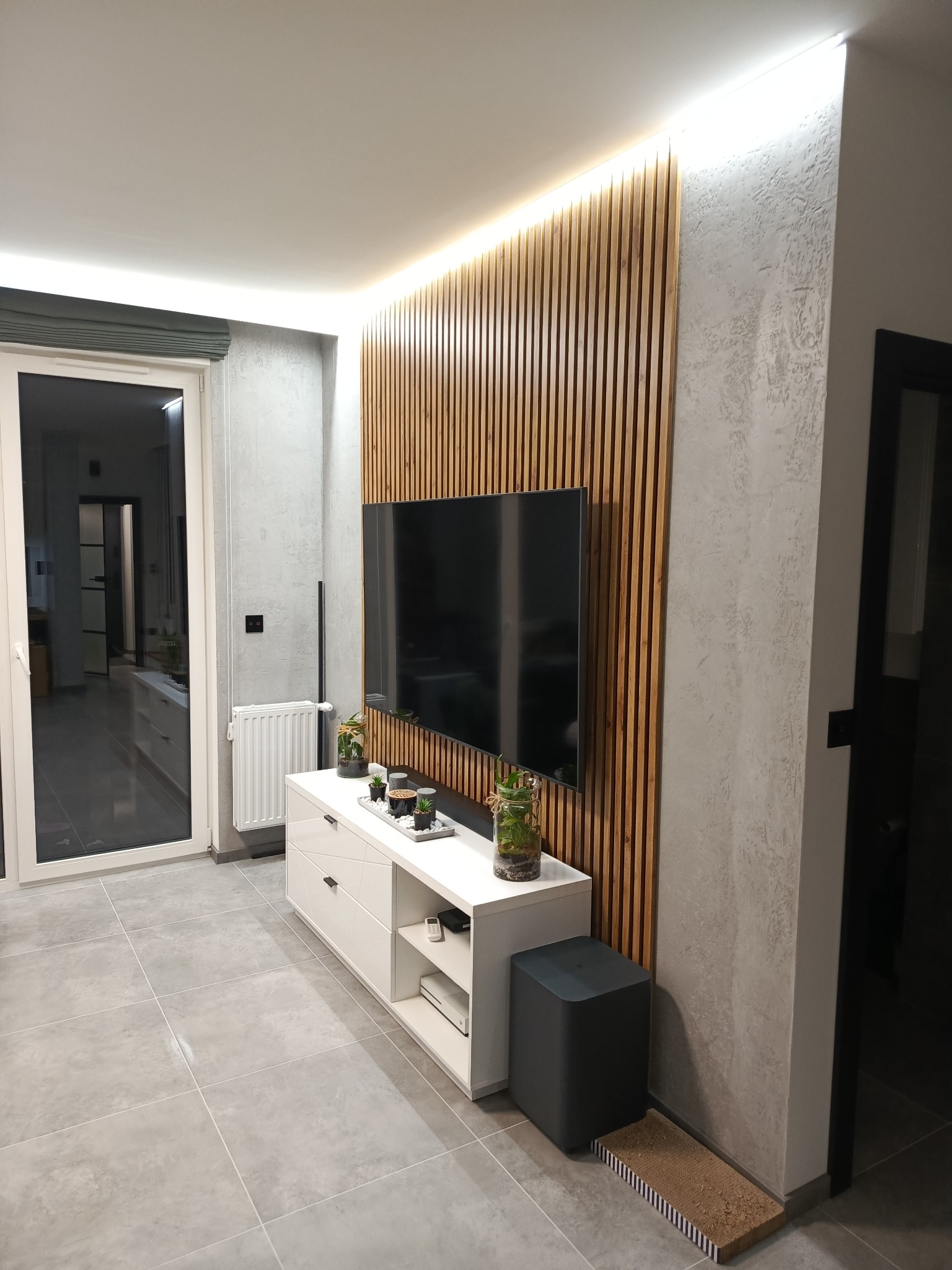 Nowoczesny salon z drewnianą ścianą, podświetleniem LED, szarą podłogą i białymi meblami. Minimalistyczny design z roślinami w szklanych naczyniach.