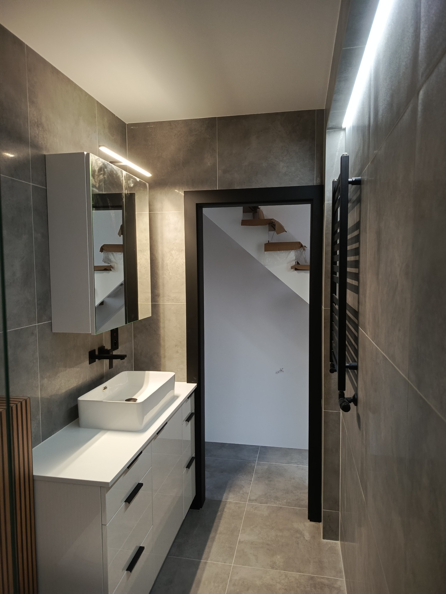 Nowoczesna łazienka z szarymi płytkami, białą szafką pod umywalką i czarnym grzejnikiem drabinkowym. W tle widoczne schody.