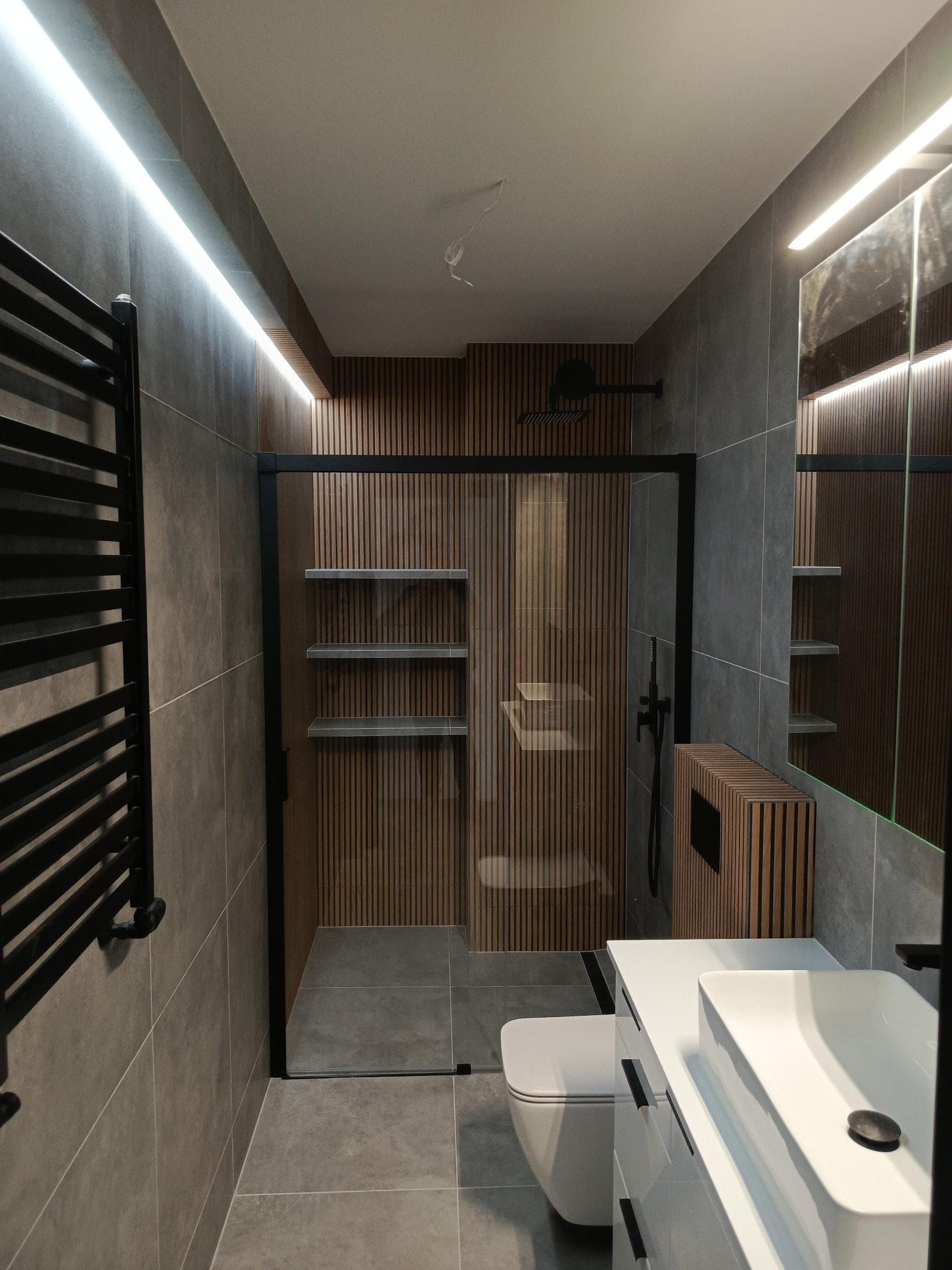 Nowoczesna łazienka z kabiną typu walk-in, wykończona w szarościach i drewnie. Grzejnik drabinkowy w kolorze czarnym. Realizacja w Starogardzie Gdańskim.