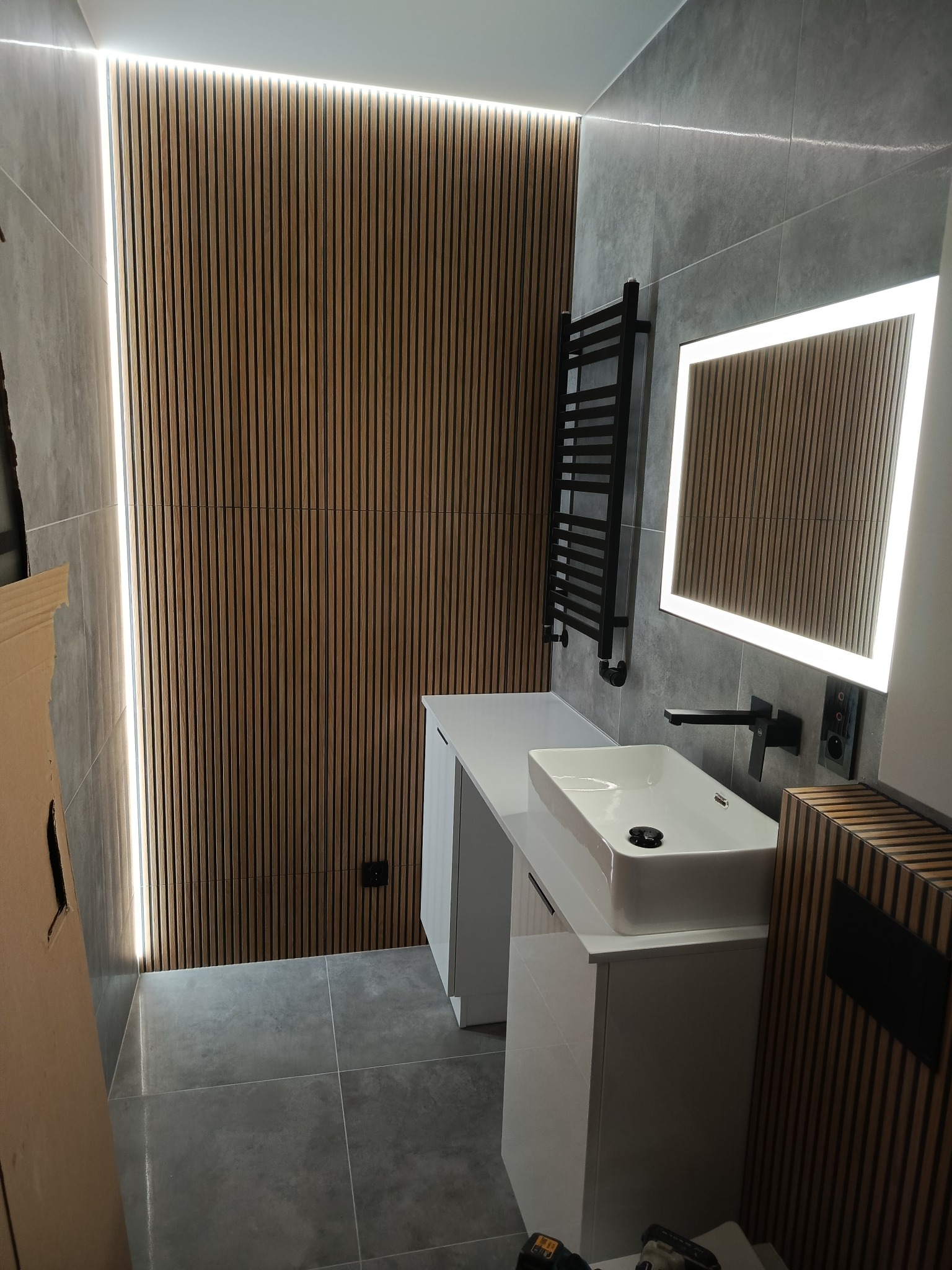 Nowoczesna łazienka z szarą płytką, drewnianymi lamelami, podświetlanym lustrem i białą umywalką nablatową. Minimalistyczny design z czarnymi akcentami.