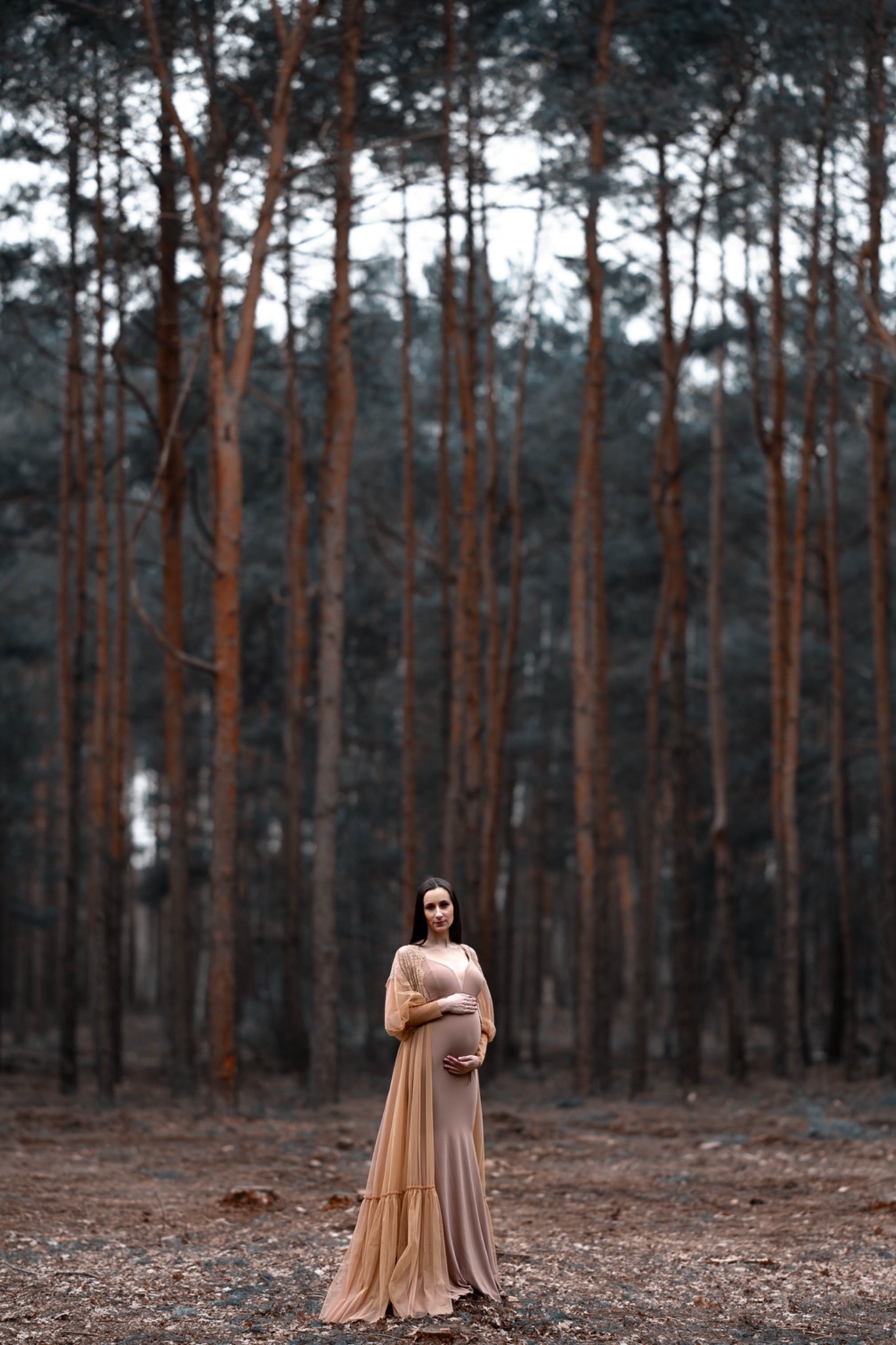 Kobieta w ciążowej sukni w kolorze beżowym z narzutką z tiulu, pozuje na tle lasu z wysokimi drzewami.