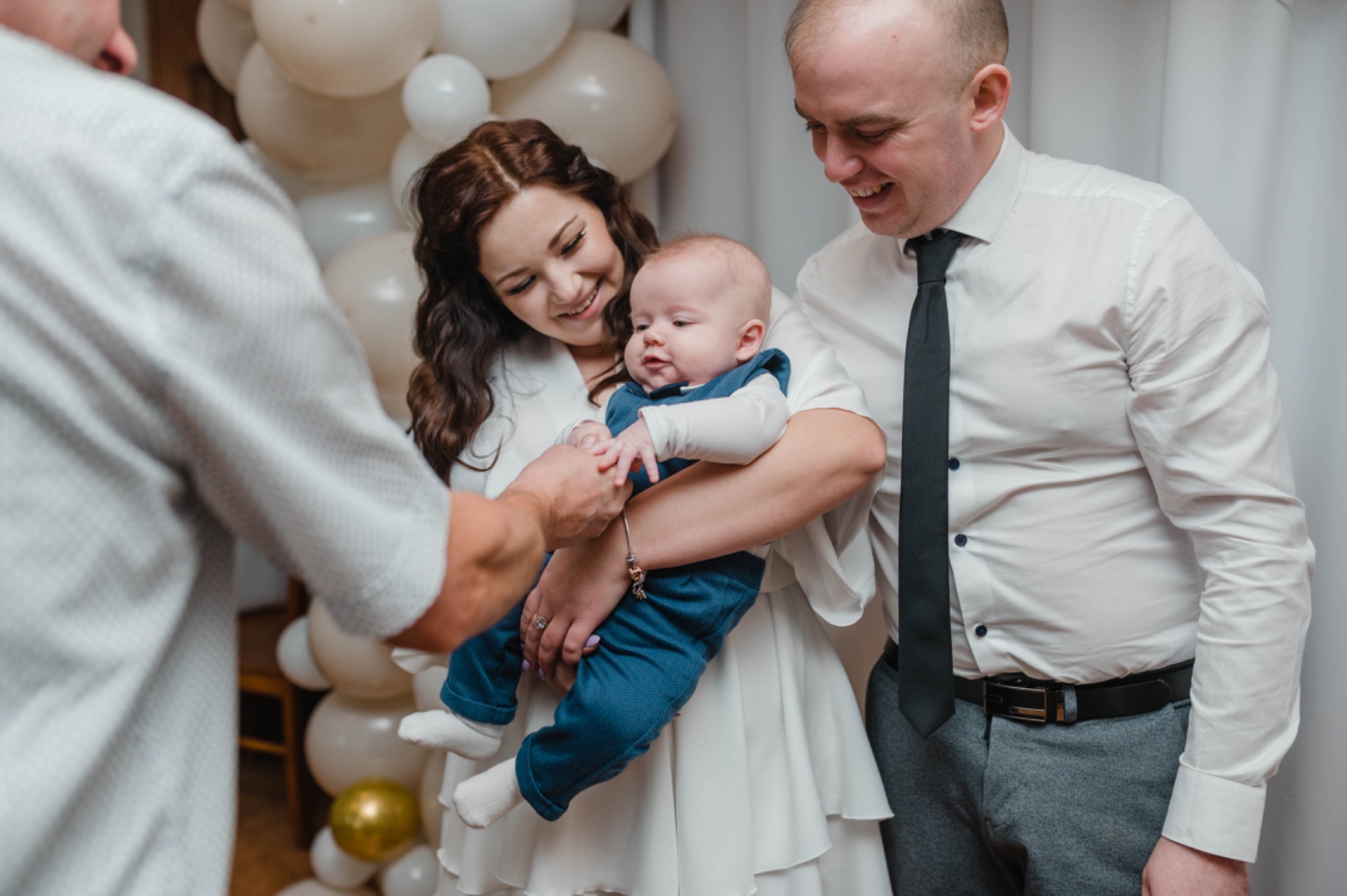 Uśmiechnięta matka trzyma niemowlę w niebieskim ubranku, ojciec z boku również się uśmiecha, a dziadek poprawia ubranko dziecka na tle białych i złotych balonów.
