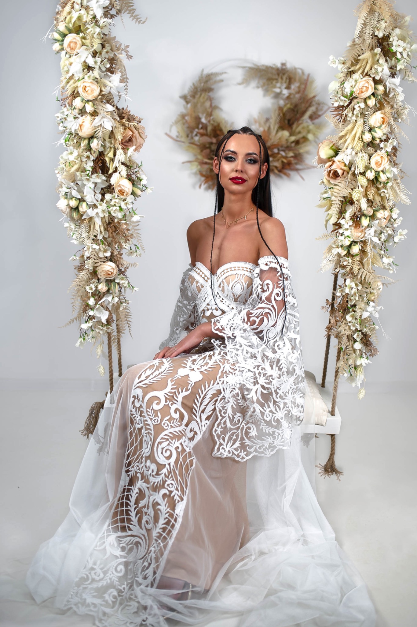 Kobieta w białej, koronkowej sukni, siedząca na huśtawce udekorowanej kwiatami, na tle białej ściany z wiankiem.