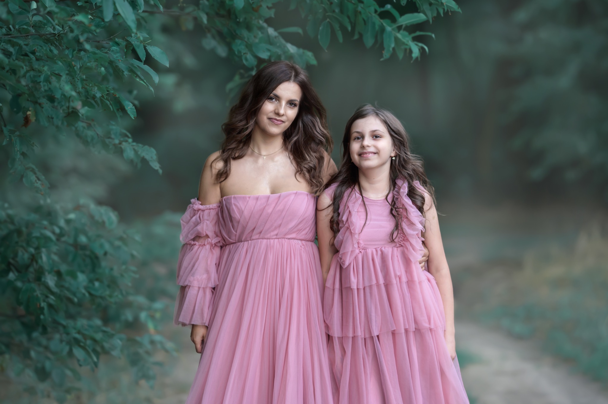 Portret matki i córki w dopasowanych, zwiewnych różowych sukniach na tle leśnej scenerii z zielonymi liśćmi i rozmytą ścieżką.