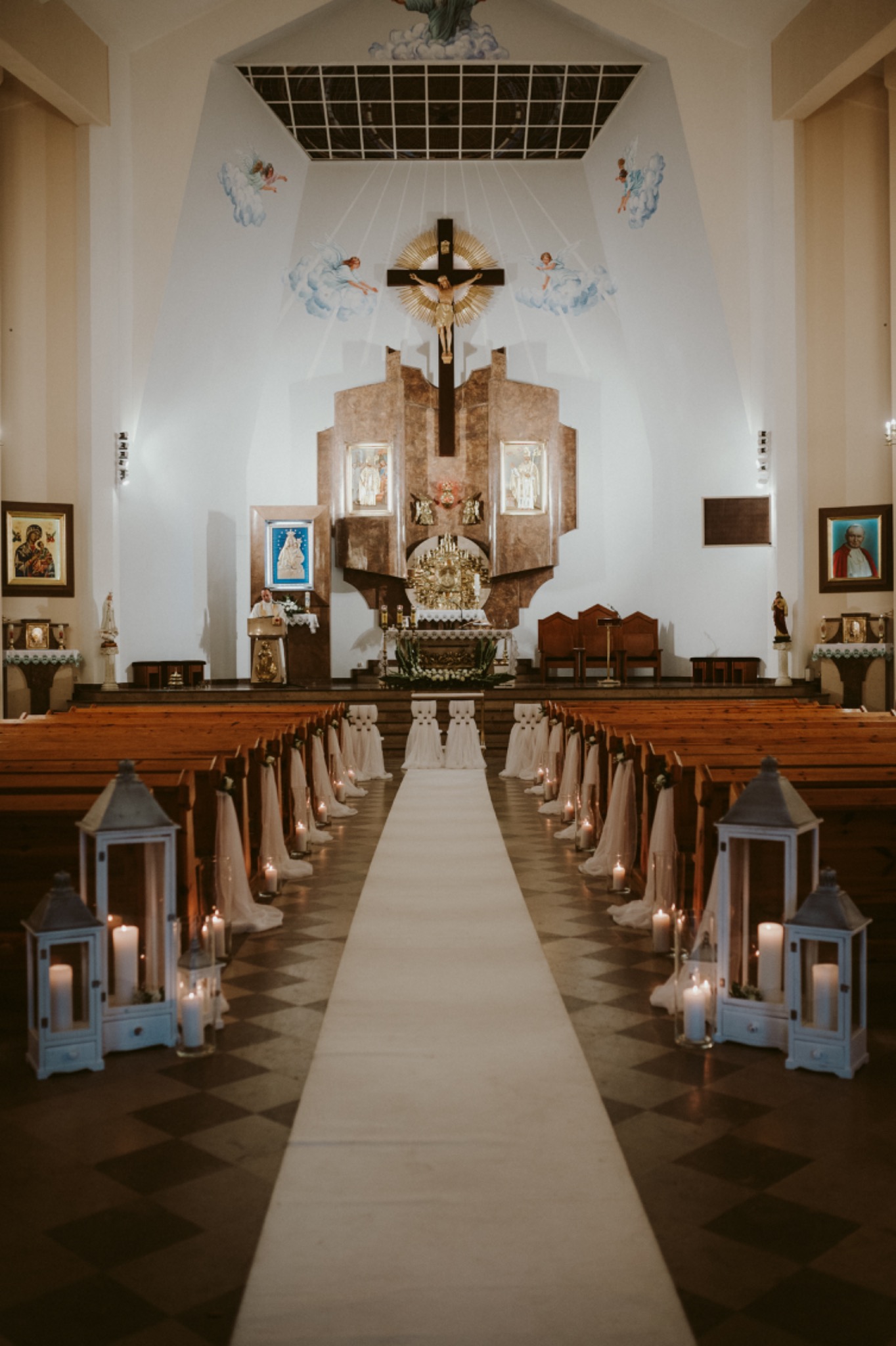 Uroczystość ślubna w kościele: biały dywan, lampiony ze świecami wzdłuż ławek, dekoracje z tiulu, ołtarz w tle.