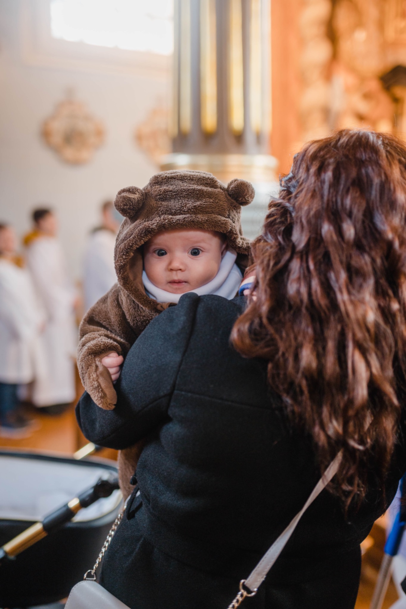 Matka trzyma na rękach niemowlę ubrane w brązowy kombinezon z kapturem imitującym misia, w tle widoczni ludzie w białych szatach i fragment wnętrza kościoła.
