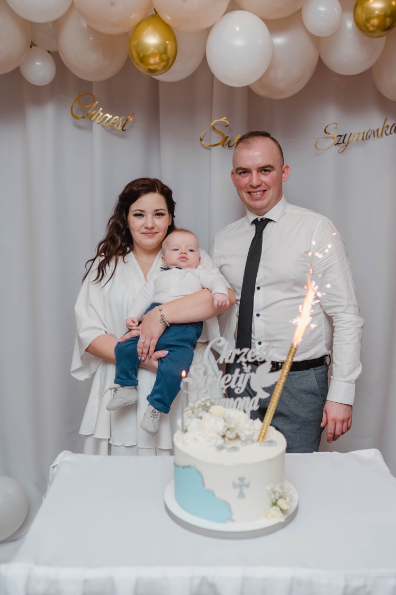 Uśmiechnięta rodzina pozuje za tortem z płonącą świeczką, dekoracjami i napisem 'Chrzest Święty Szymona' na tle białych balonów i zasłon.